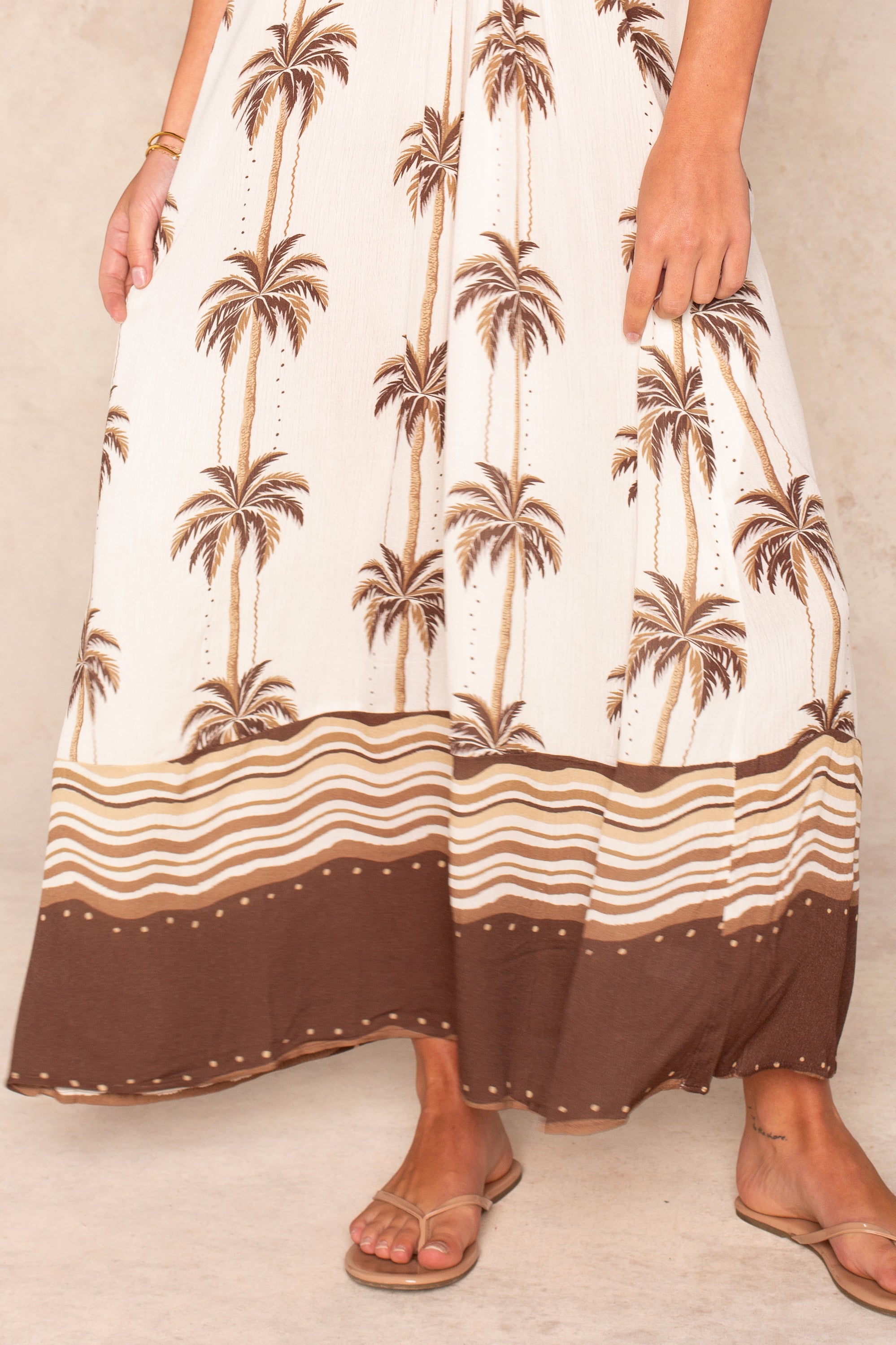 A person wearing the Isla Pauli Maxi O-ring Coverup with brown palm tree prints and a wavy striped hem, paired with tan sandals. Only the lower half of this chic beach dress is visible.