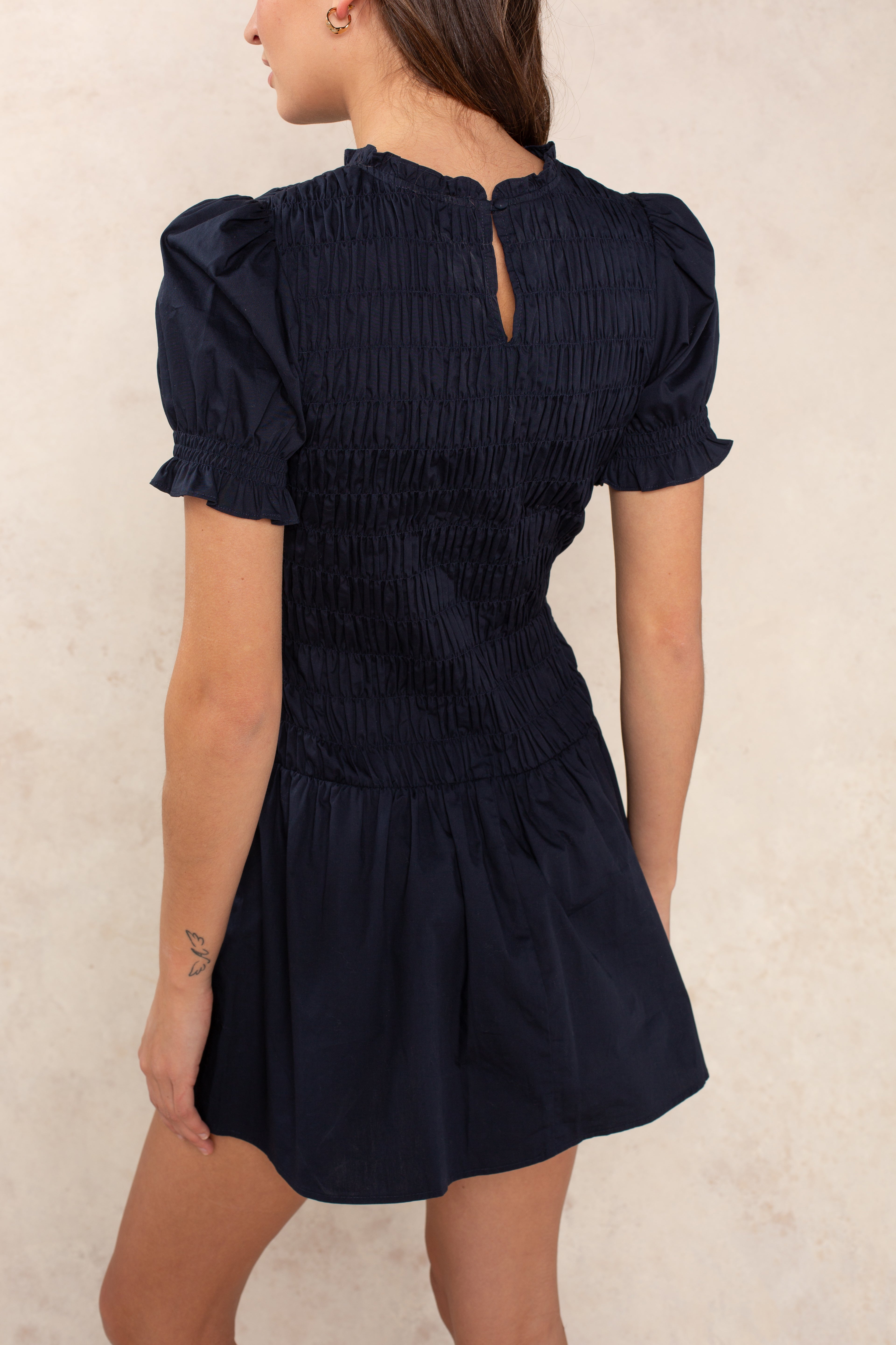 A woman with long brown hair and a small left arm tattoo stands sideways, showcasing the back of the Georgie Esme Smocked Short Sleeve Mini Dress in navy, featuring a smocked bodice, puffed sleeves, and a keyhole button neckline.