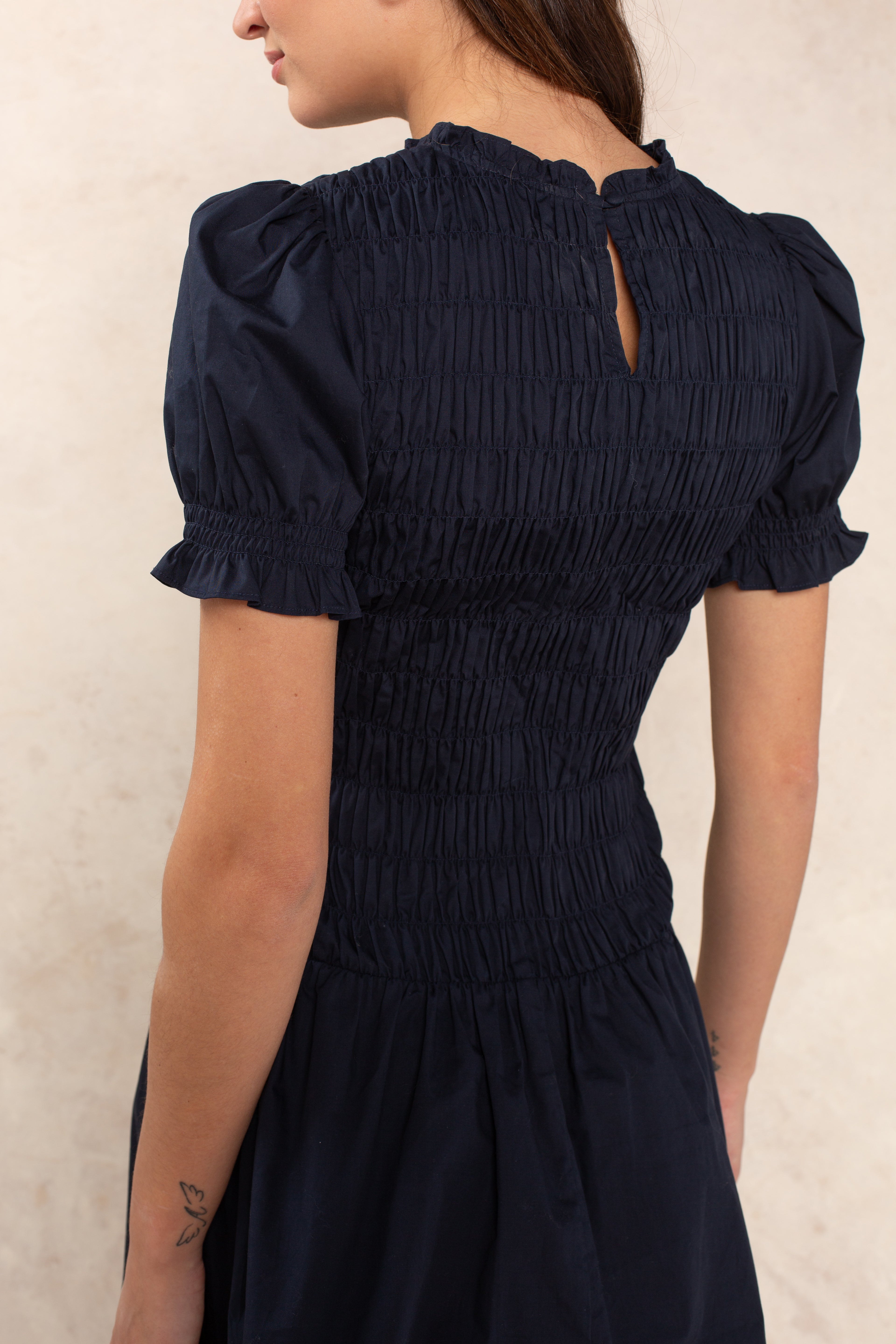 A woman with brown hair tied back is shown from the side and back wearing the Georgie Esme Smocked Short Sleeve Mini Dress in navy with a smocked bodice, ruffled cuffs, puffed sleeves, and a keyhole neckline.