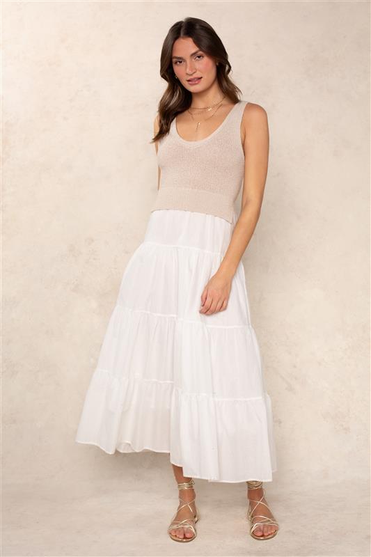 A woman poses against a light beige background wearing the Georgie Aries Dress, featuring a relaxed fit, sleeveless design in beige with tiered white skirt detail. She pairs it with minimal jewelry and strappy sandals for an effortlessly chic look.