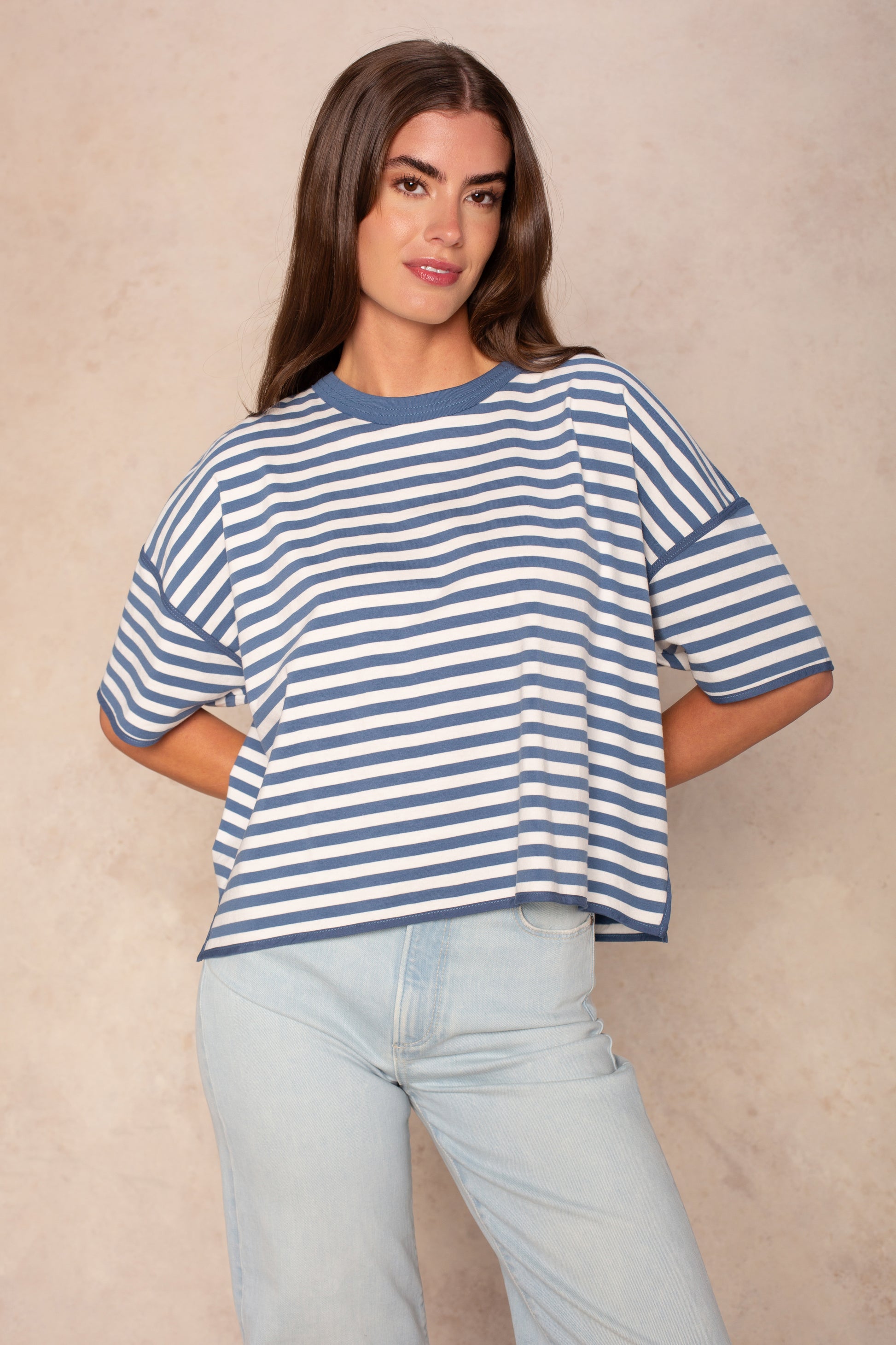 A woman with long brown hair models the Isla Franki Boxy Stripe Knit Tee—an oversized blue and white striped top—paired with light blue jeans, standing against a neutral beige background for effortless casual style.