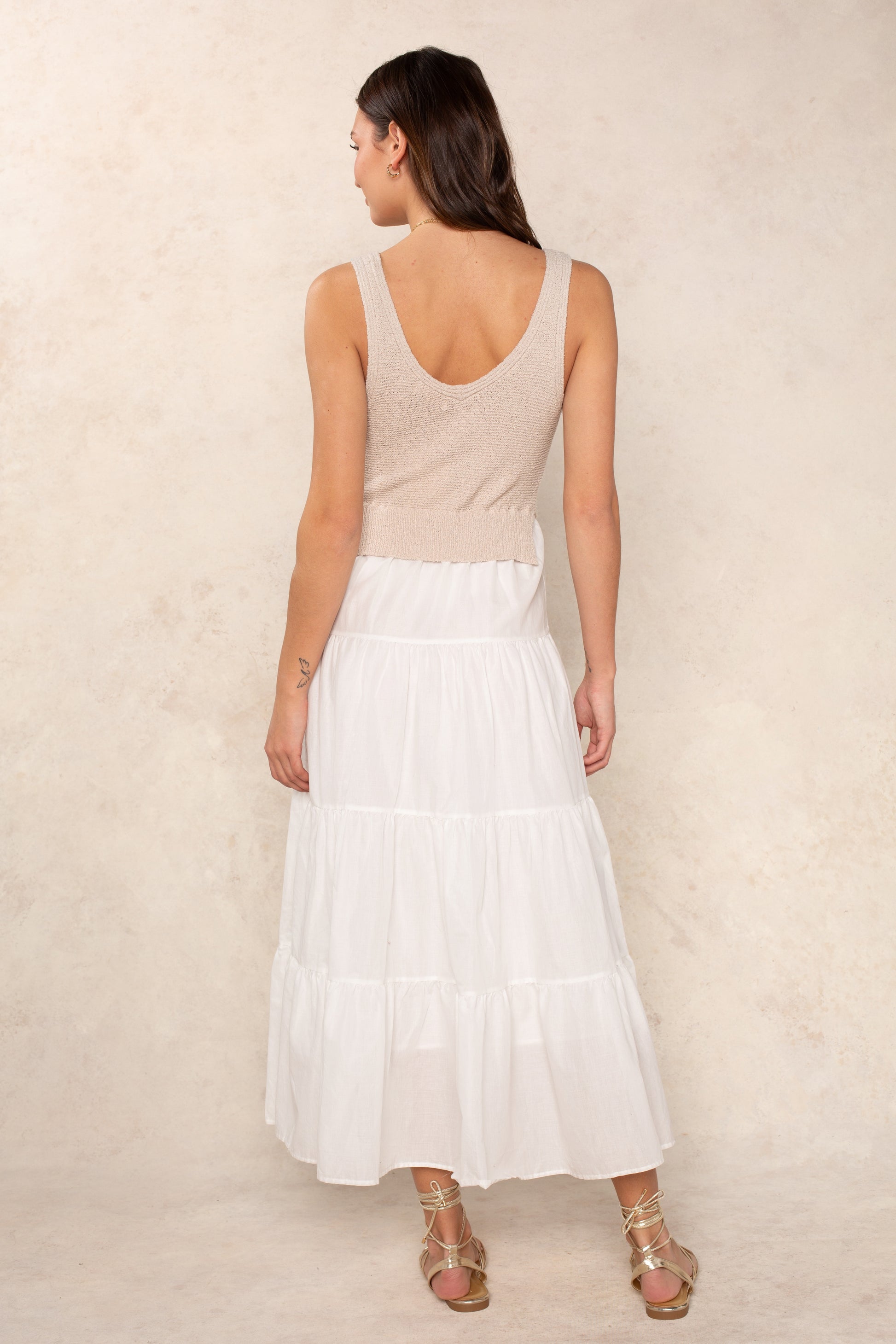 A woman stands with her back to the camera against a light textured background, wearing the Georgie Aries Dress—a beige relaxed fit sleeveless knit top and long white tiered skirt—paired with gold strappy sandals.