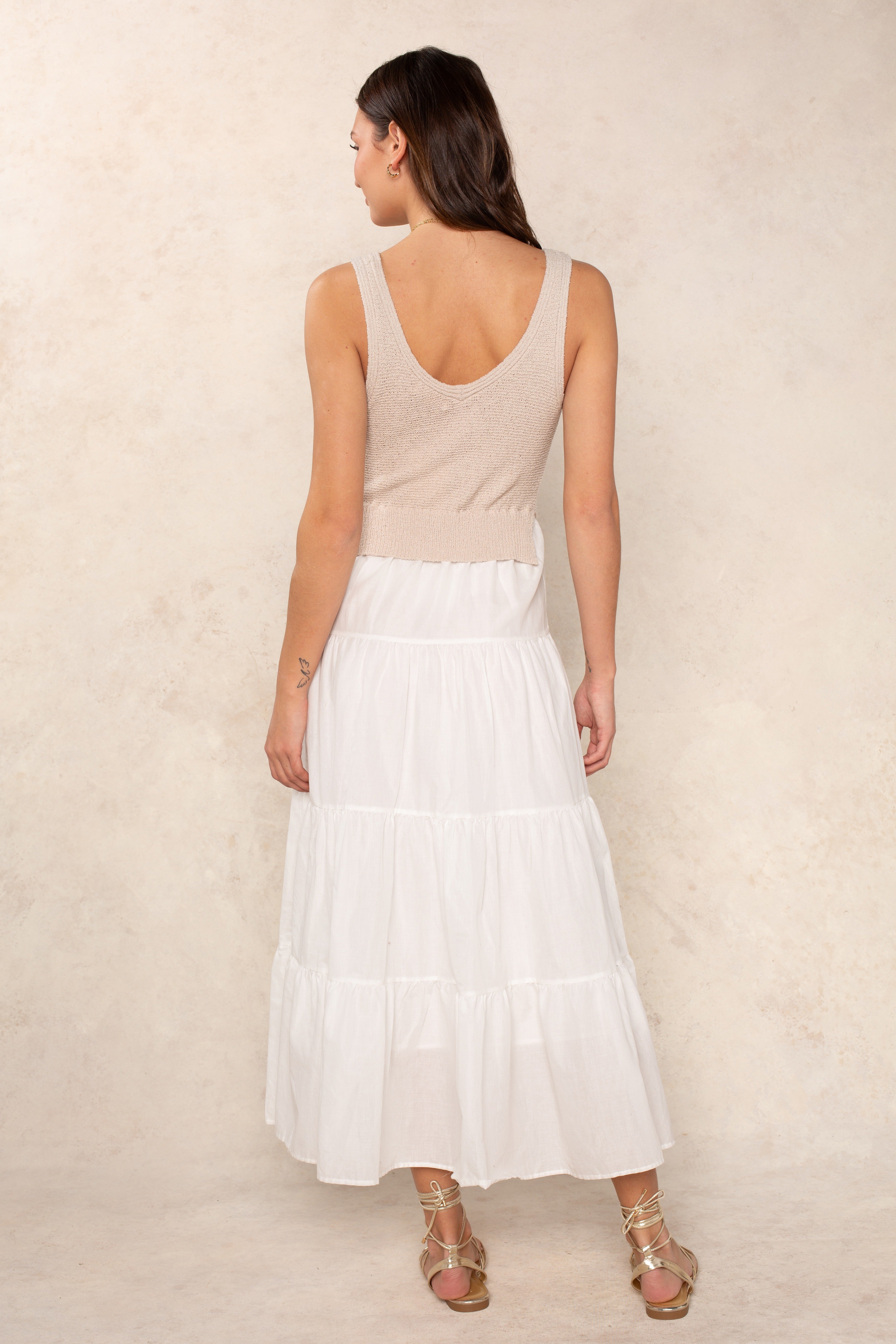 A woman stands with her back to the camera against a light textured background, wearing the Georgie Aries Dress—a beige relaxed fit sleeveless knit top and long white tiered skirt—paired with gold strappy sandals.
