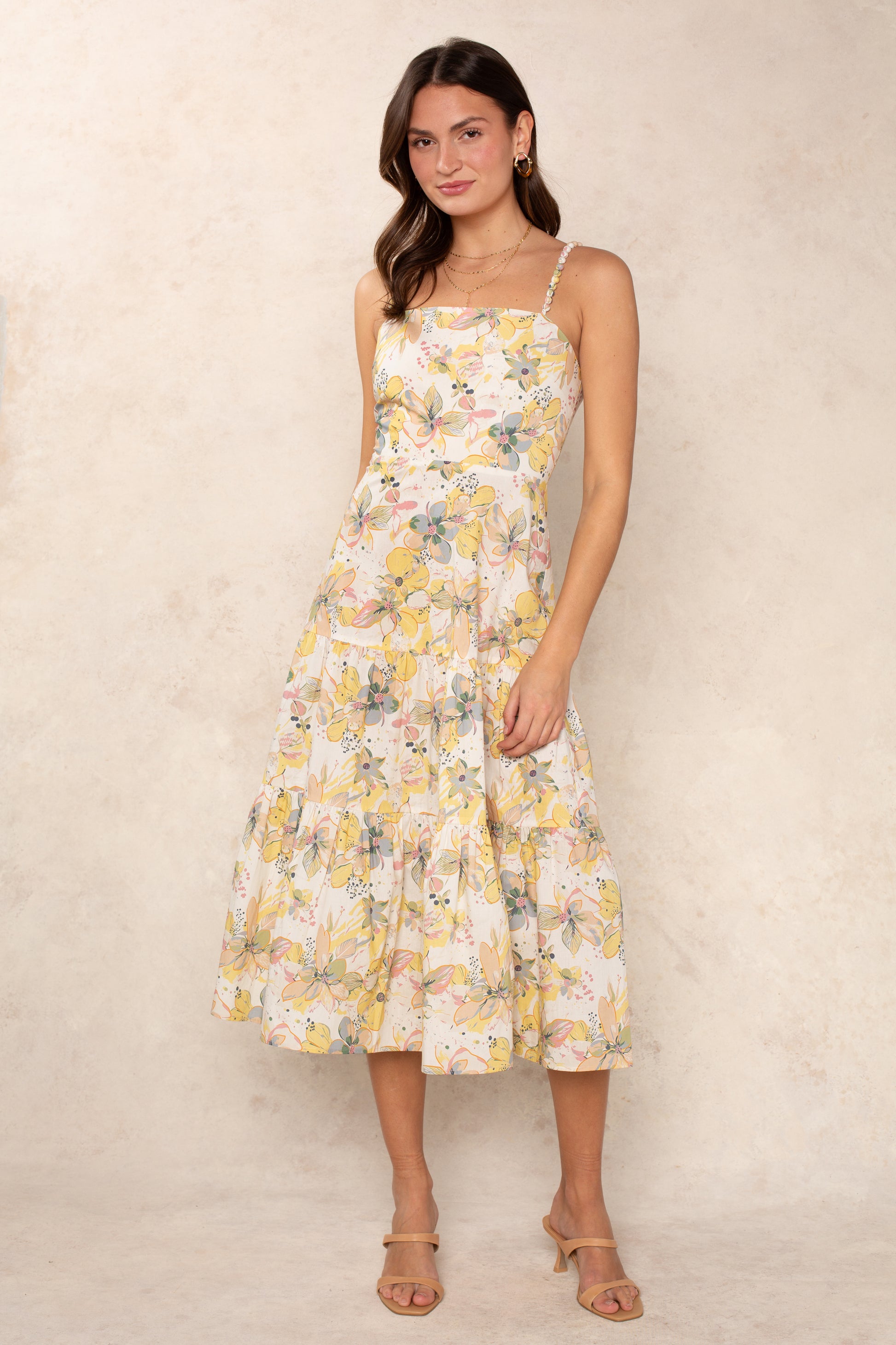 A woman poses against a light beige background in the Georgie Dagney Cotton Midi Dress, featuring a corset-style bodice, thin straps, and a tiered skirt. She completes the look with nude heeled sandals, subtle jewelry, and loose waves.