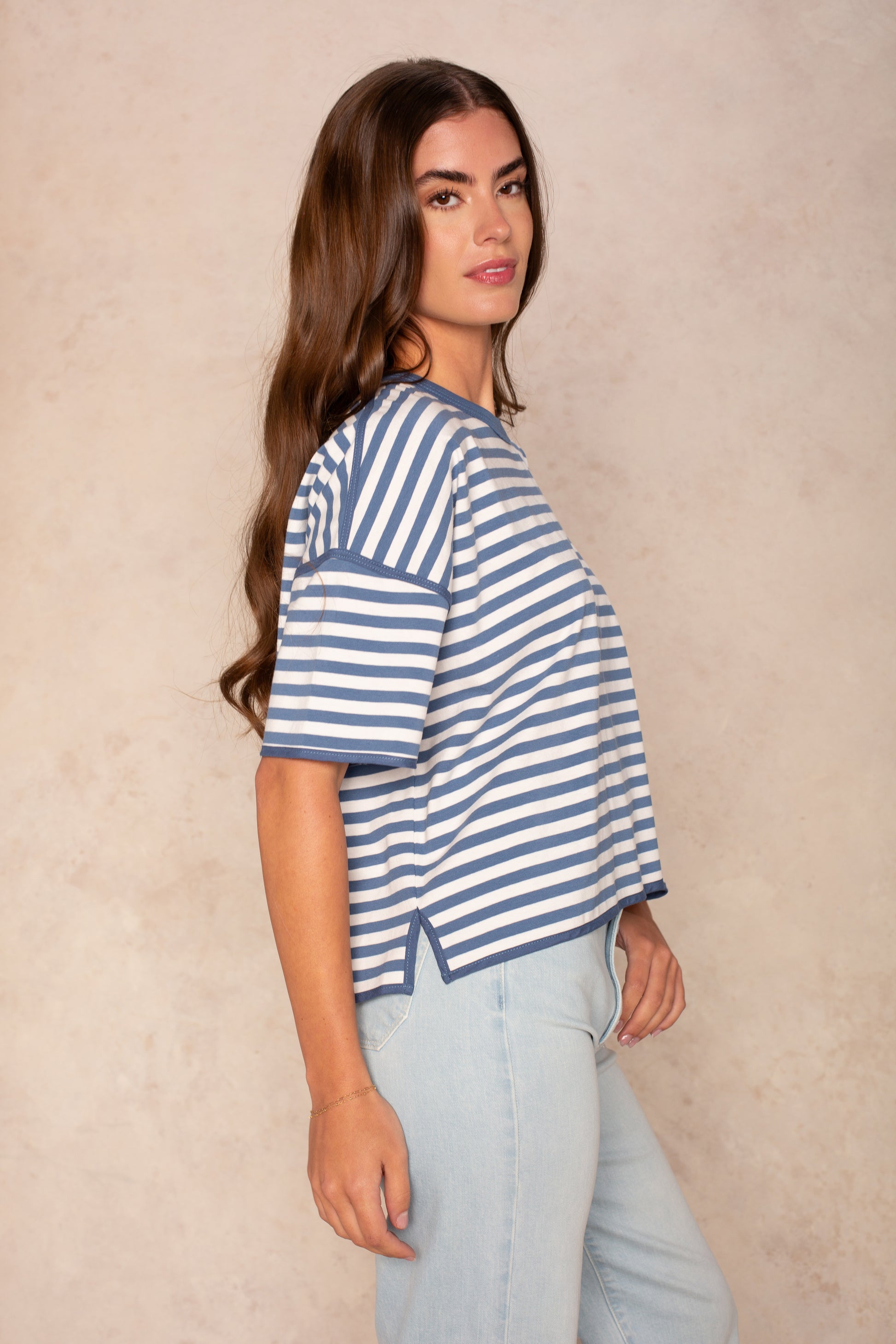 A woman with long brown hair stands in profile against a beige background, wearing the Isla Franki Boxy Stripe Knit Tee in blue and white stripes and light blue jeans for a relaxed, casual look.