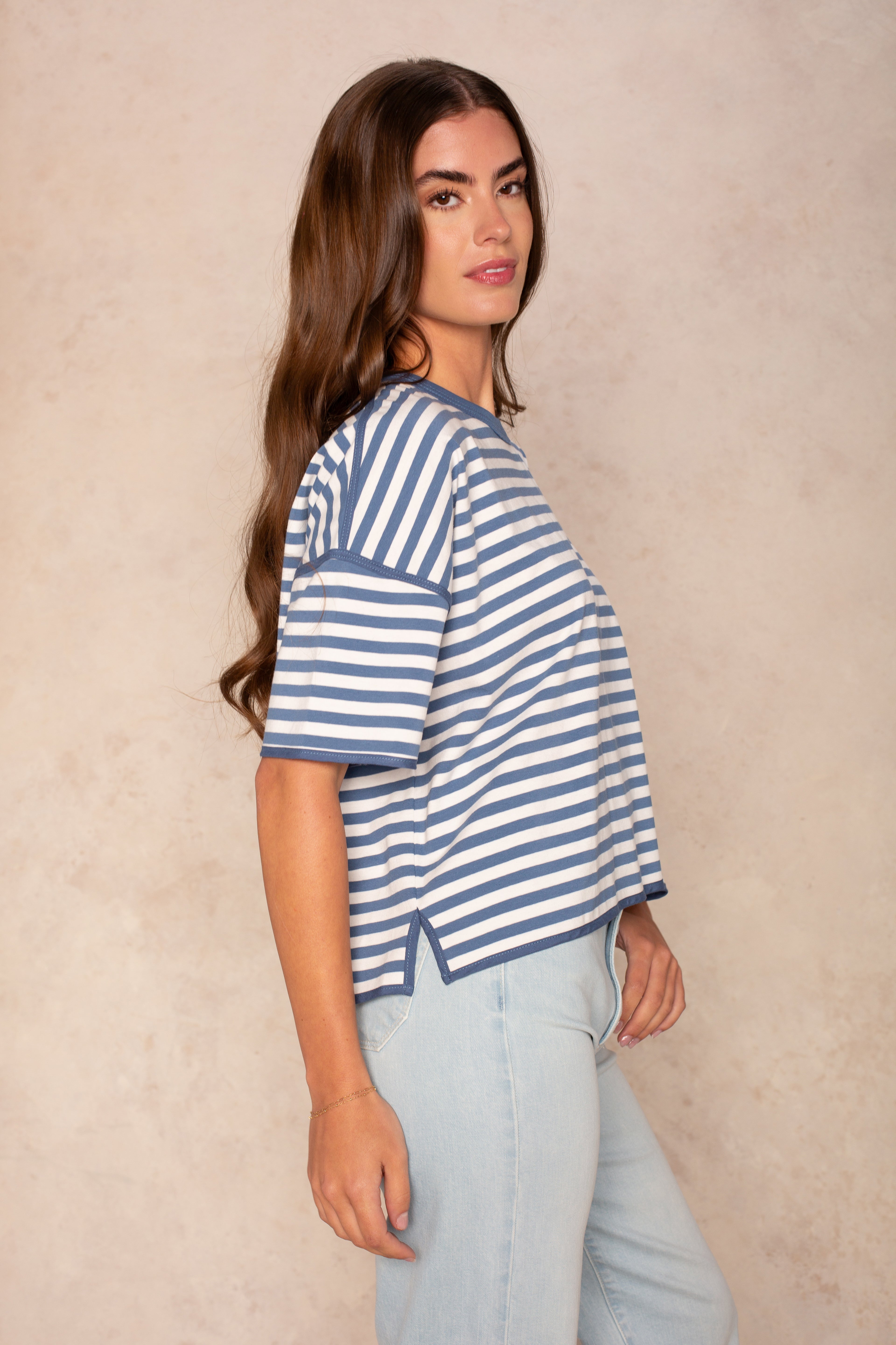 A woman with long brown hair stands in profile against a beige background, wearing the Isla Franki Boxy Stripe Knit Tee in blue and white stripes and light blue jeans for a relaxed, casual look.