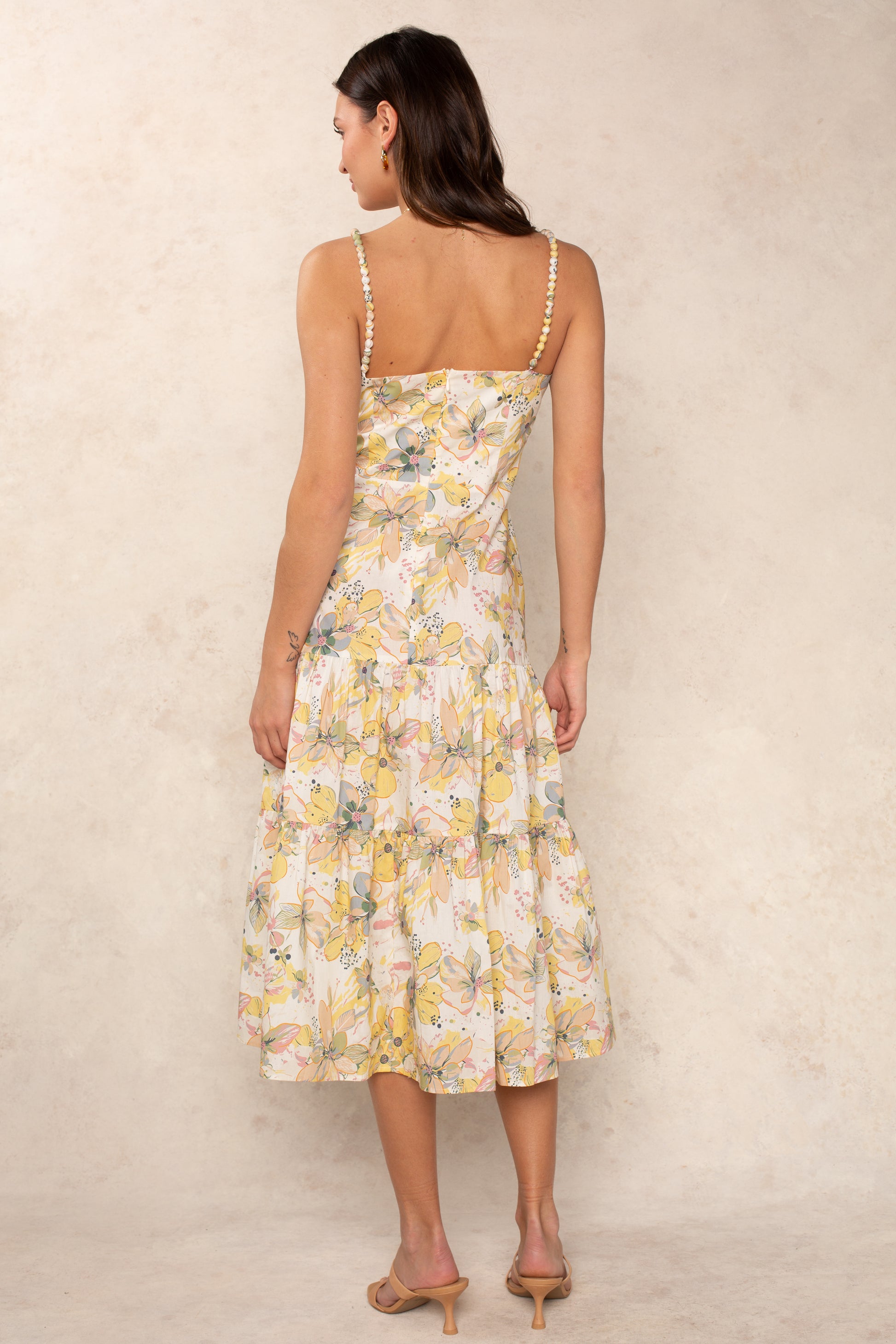 A woman faces away, wearing the Georgie Dagney Cotton Midi Dress featuring a sleeveless design, square neckline, pastel floral print, and pearl straps. The flowing ankle-length skirt is paired with beige heeled sandals against a neutral backdrop.