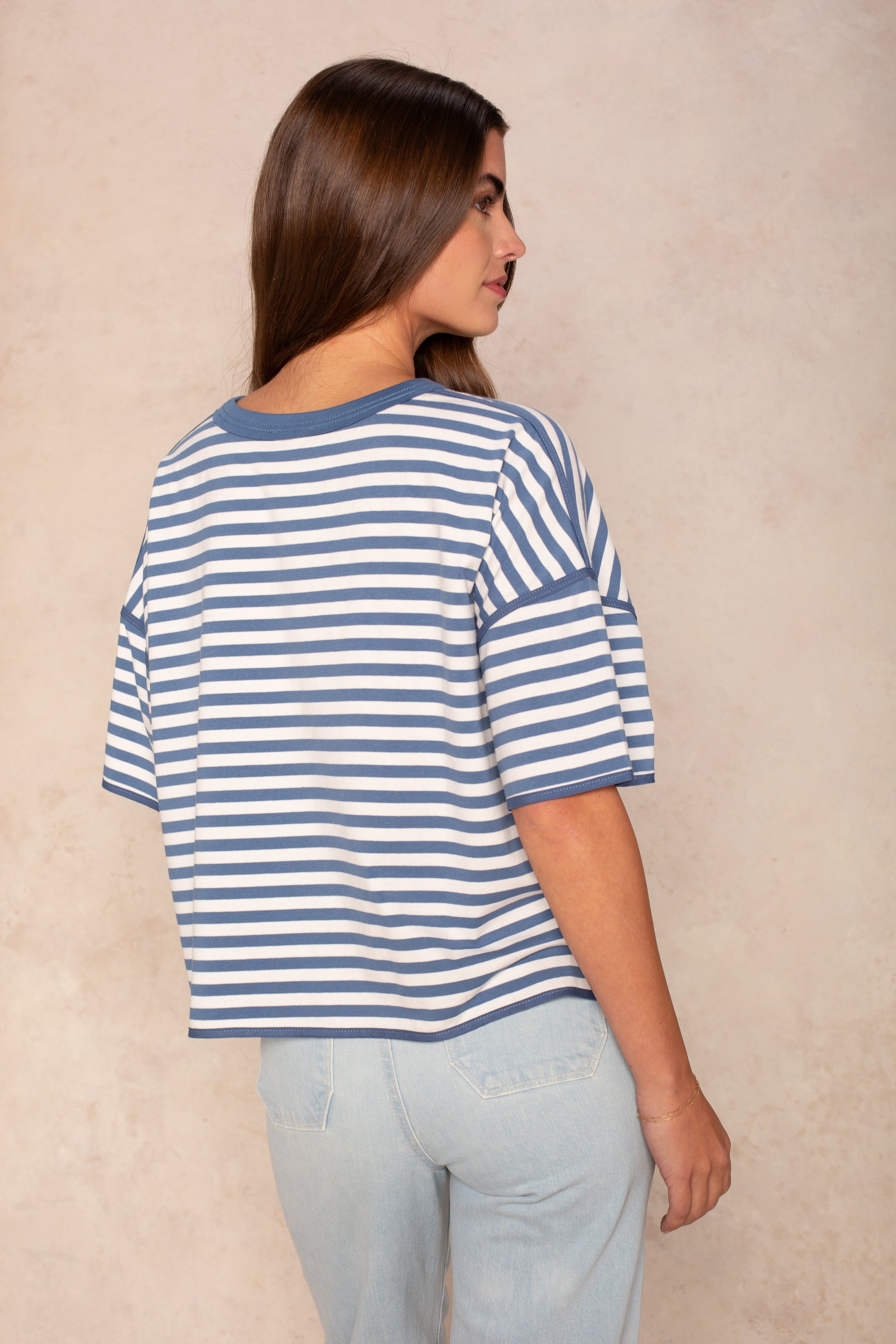 A woman with long brown hair models the Isla Franki Boxy Stripe Knit Tee in blue and white stripes with light jeans, facing away from the camera and turning her head slightly, set against a beige background.