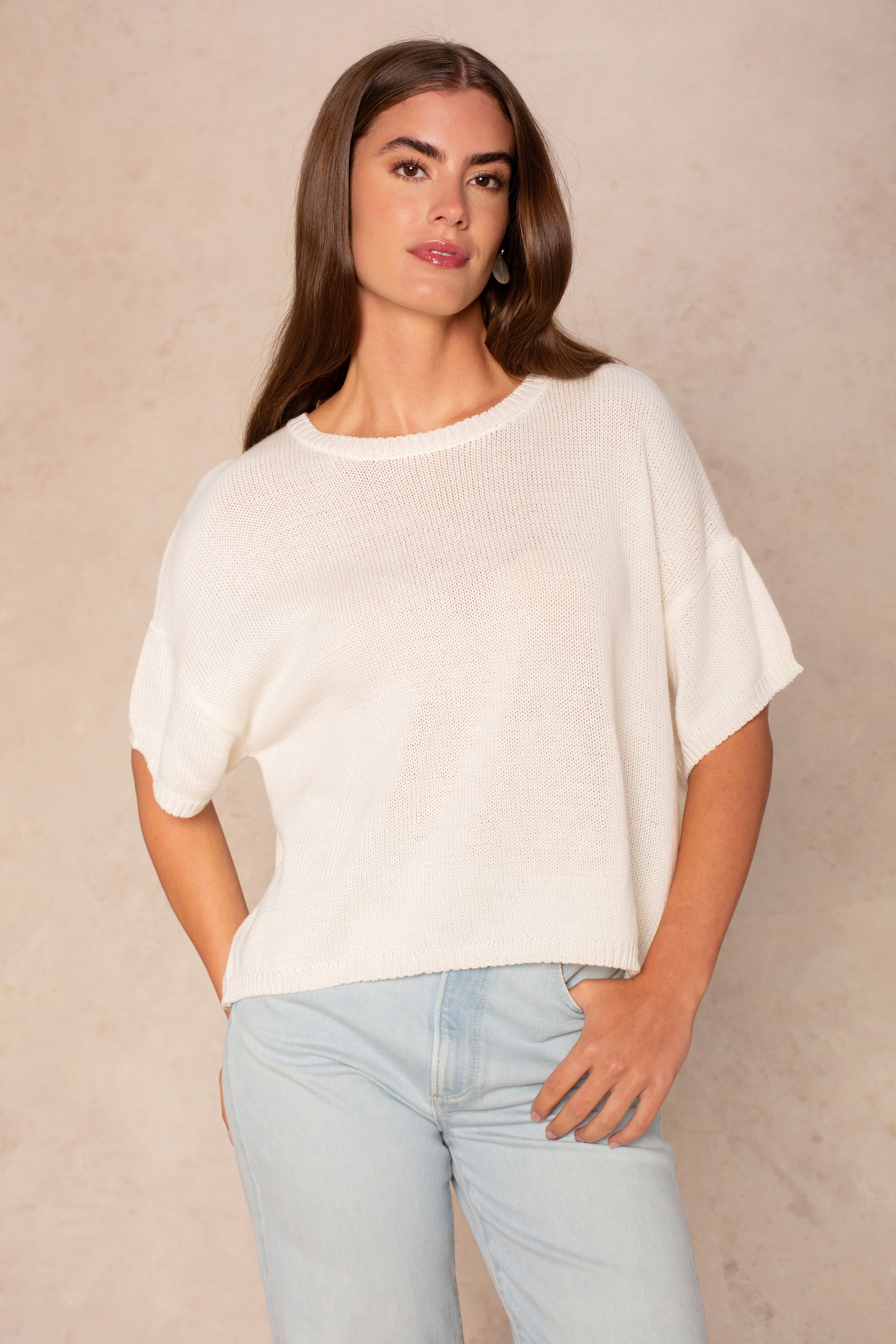 A woman with long brown hair wears the Isla Lola Square Body, an oversized white short-sleeve knit sweater, paired with light blue jeans and standing against a neutral beige background with one hand in her pocket.