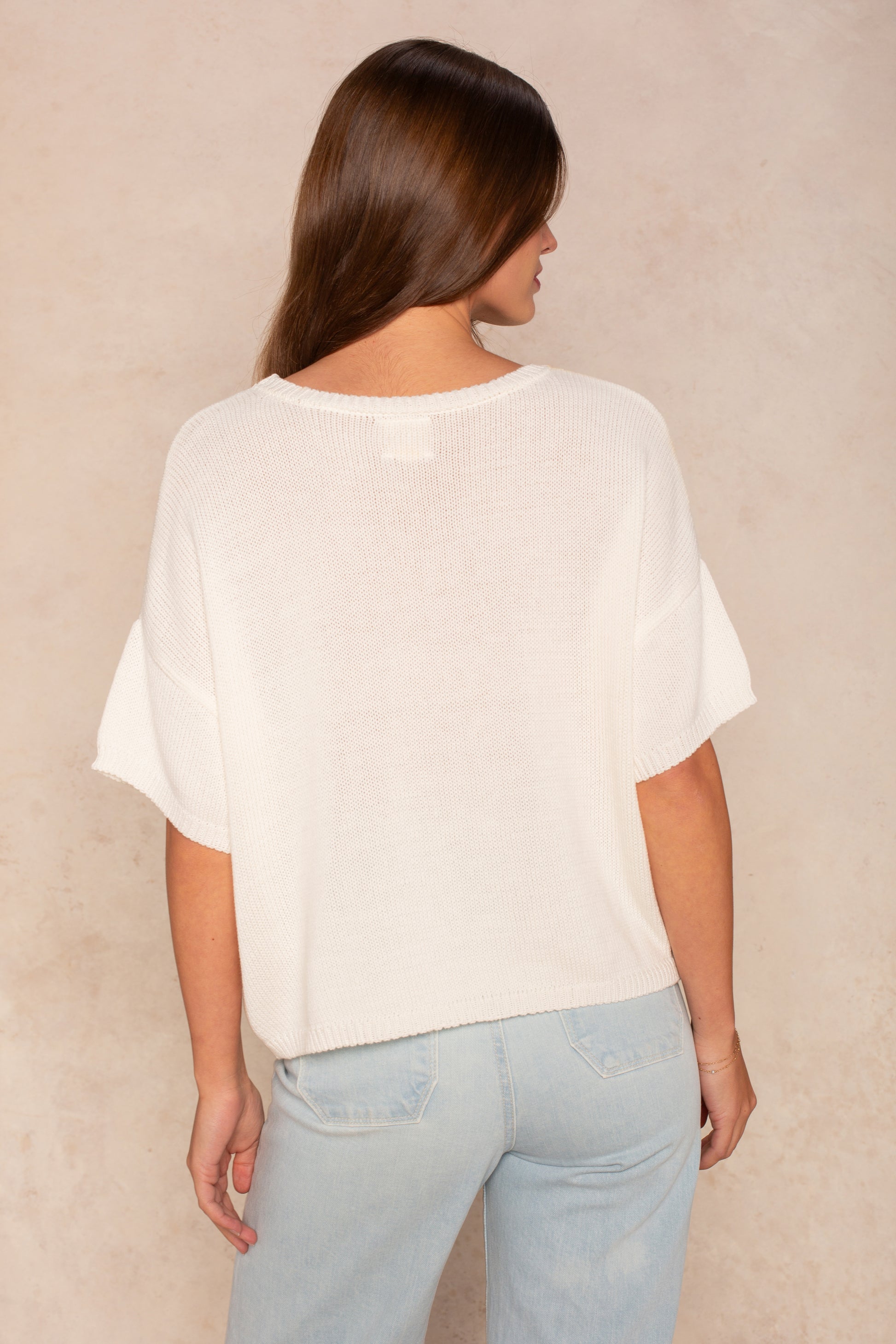 A woman with long brown hair faces away from the camera, wearing Isla's Lola Square Body oversized white short-sleeve knit top and light blue jeans, creating a casual look against a neutral beige background.