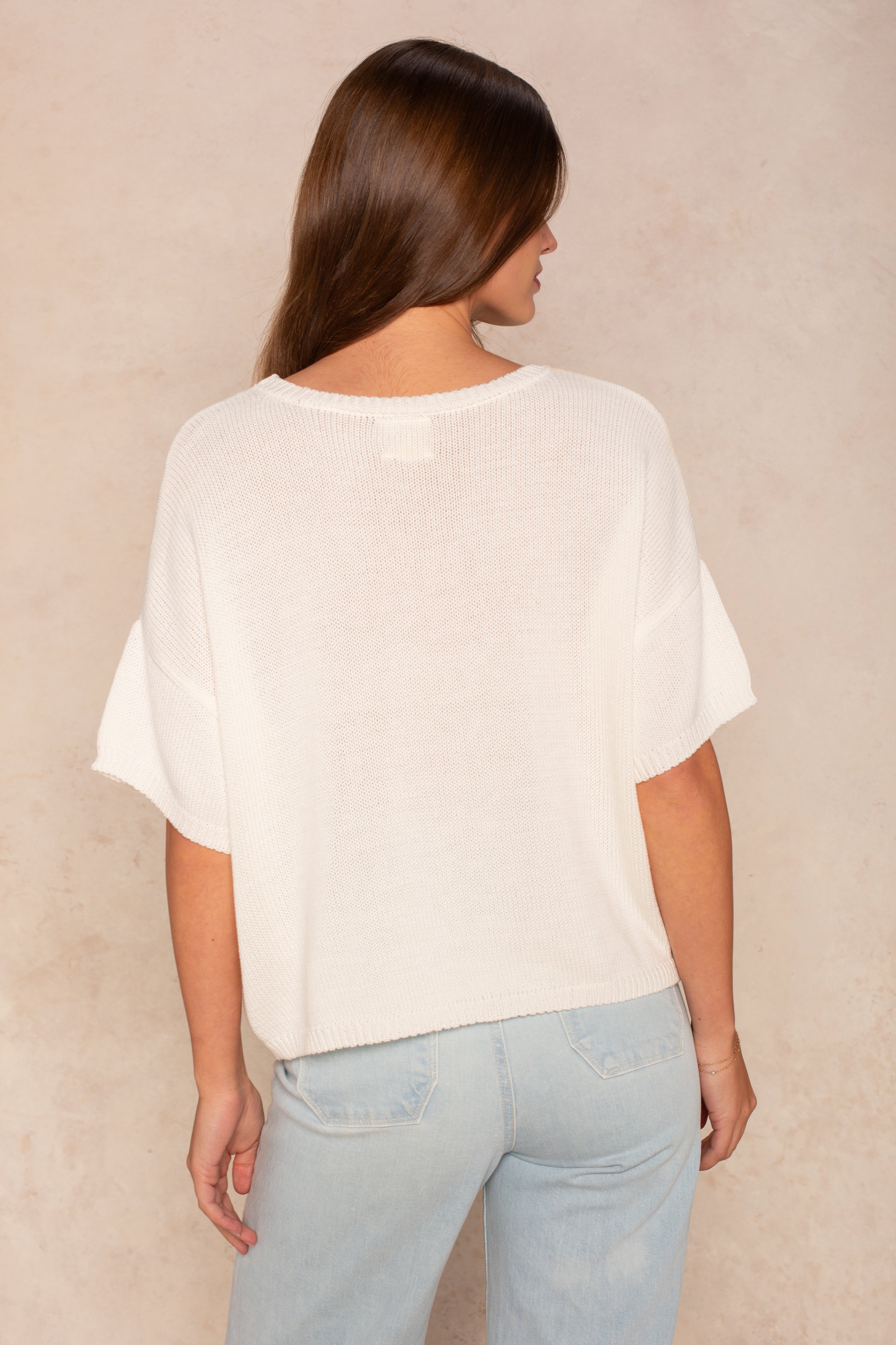 A woman with long brown hair faces away from the camera, wearing Isla's Lola Square Body oversized white short-sleeve knit top and light blue jeans, creating a casual look against a neutral beige background.