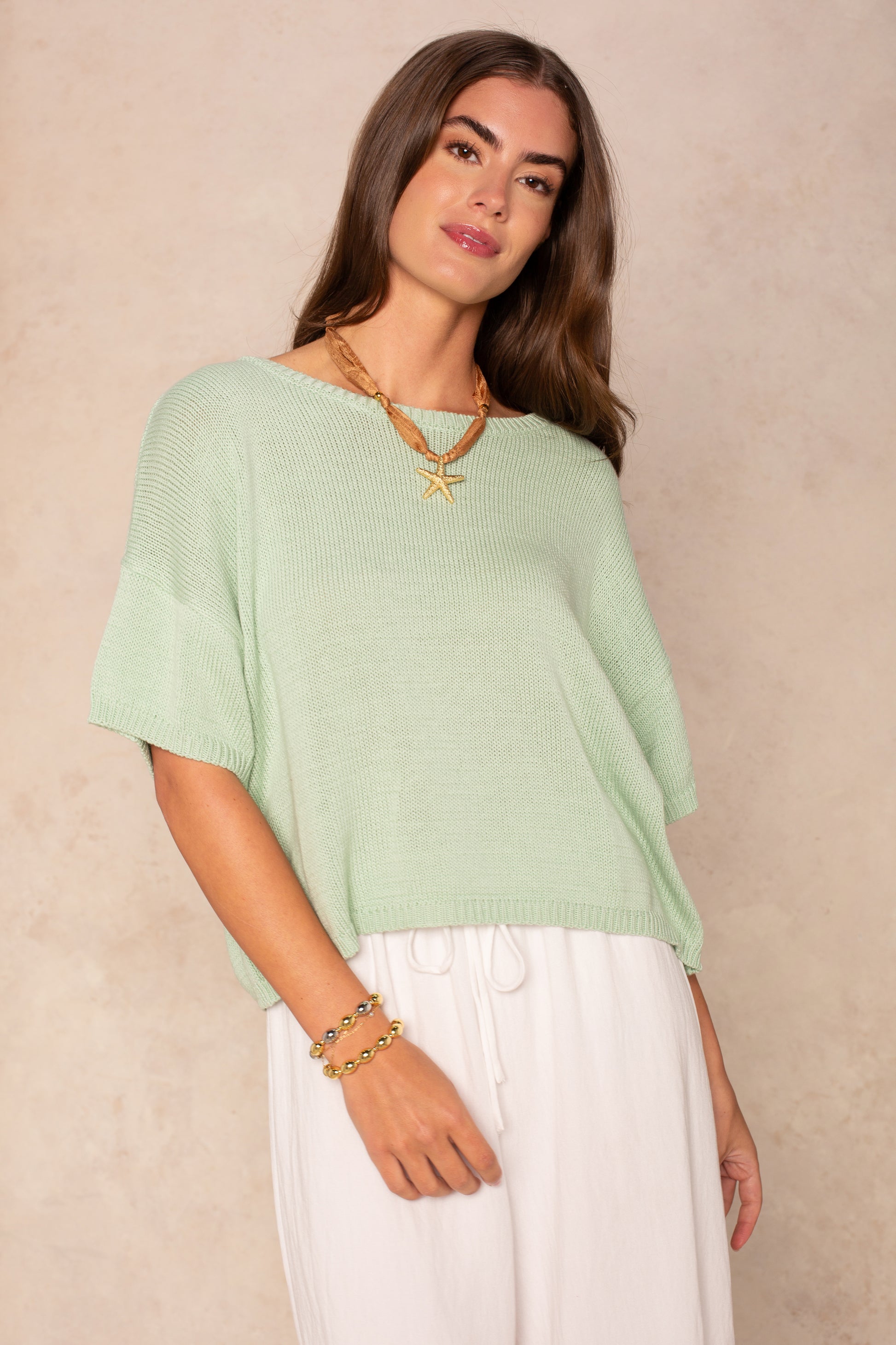 A woman with long brown hair models the Isla Lola Square Body, a loose light green short-sleeve knit top in an oversized fit, paired with white pants and gold jewelry, standing against a beige background for a casual look.