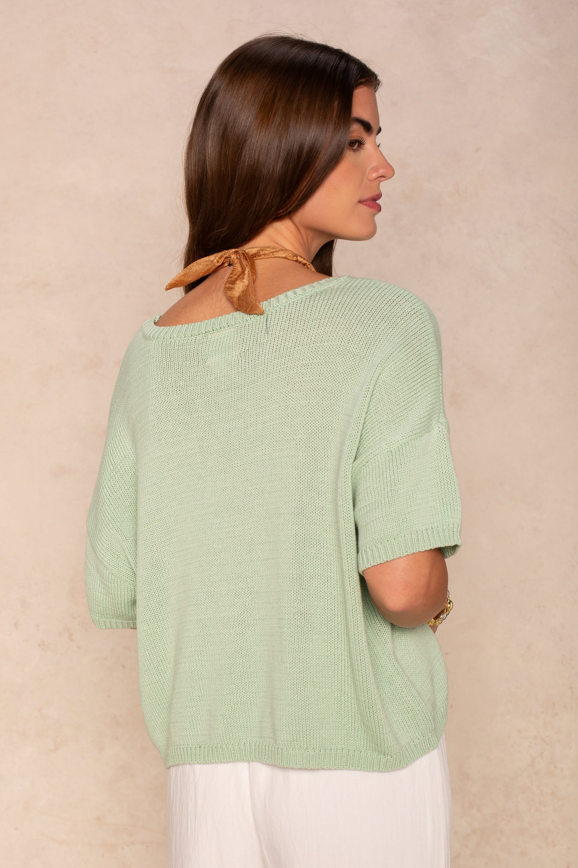 A woman with long brown hair wears the Isla Lola Square Body in light green, a short-sleeve lightweight knit top, paired with white pants, standing sideways and looking over her shoulder against a beige background.