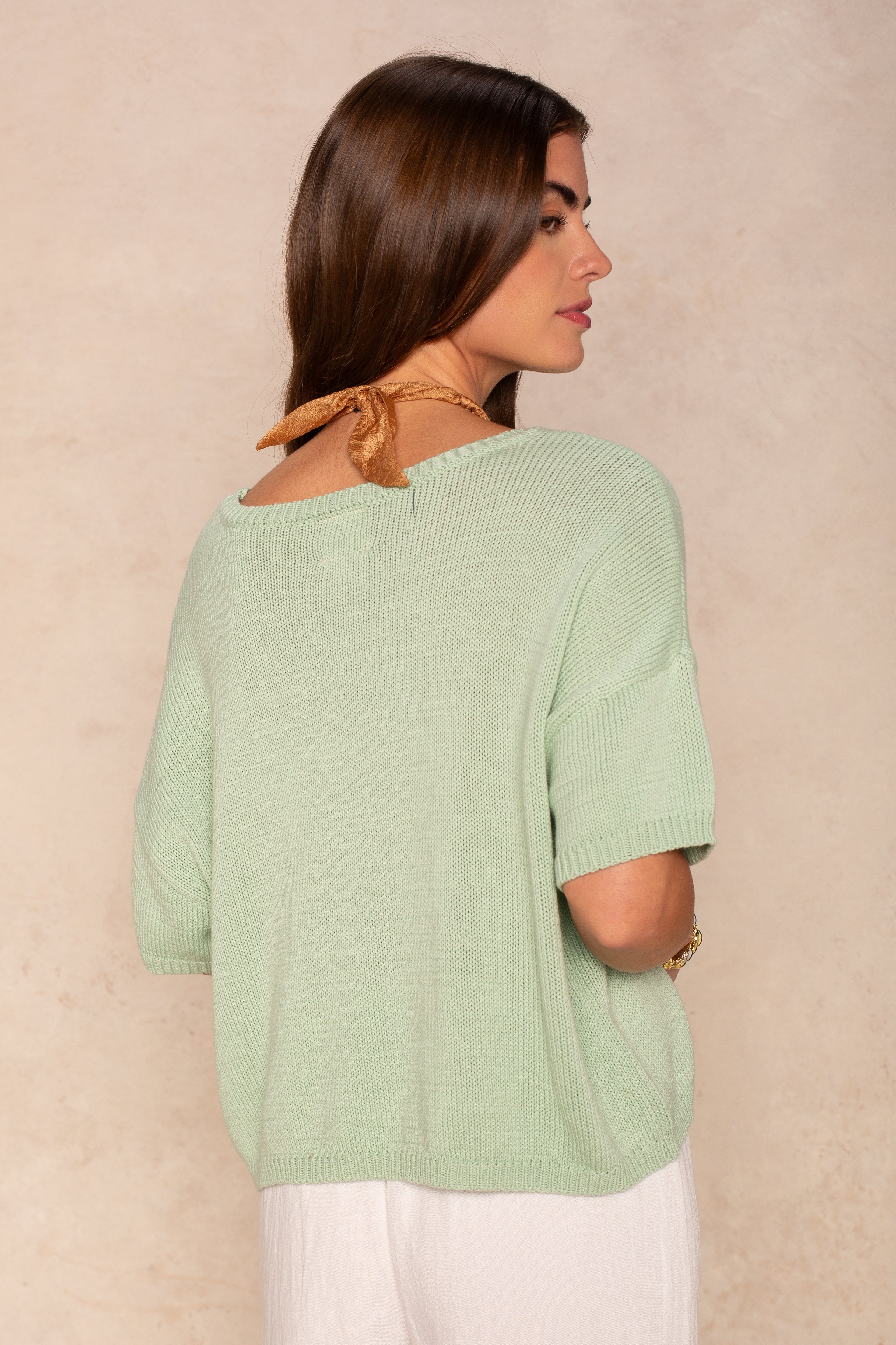 A woman with long brown hair wears the Isla Lola Square Body in light green, a short-sleeve lightweight knit top, paired with white pants, standing sideways and looking over her shoulder against a beige background.