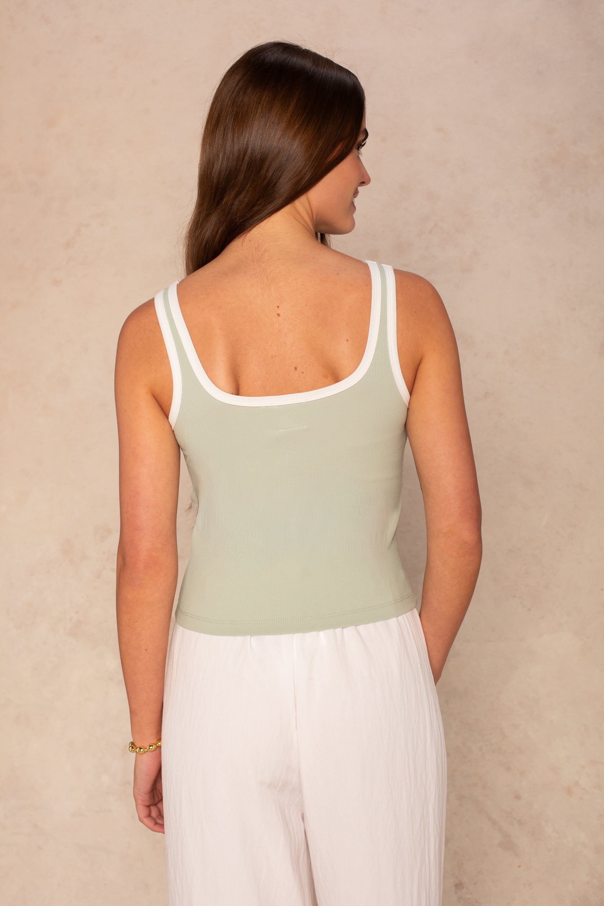 A woman with long brown hair faces away, wearing the Isla Maggie Two Tone Tank—a sleeveless, soft knit light green top with white trim—paired with loose white pants against a neutral textured background.