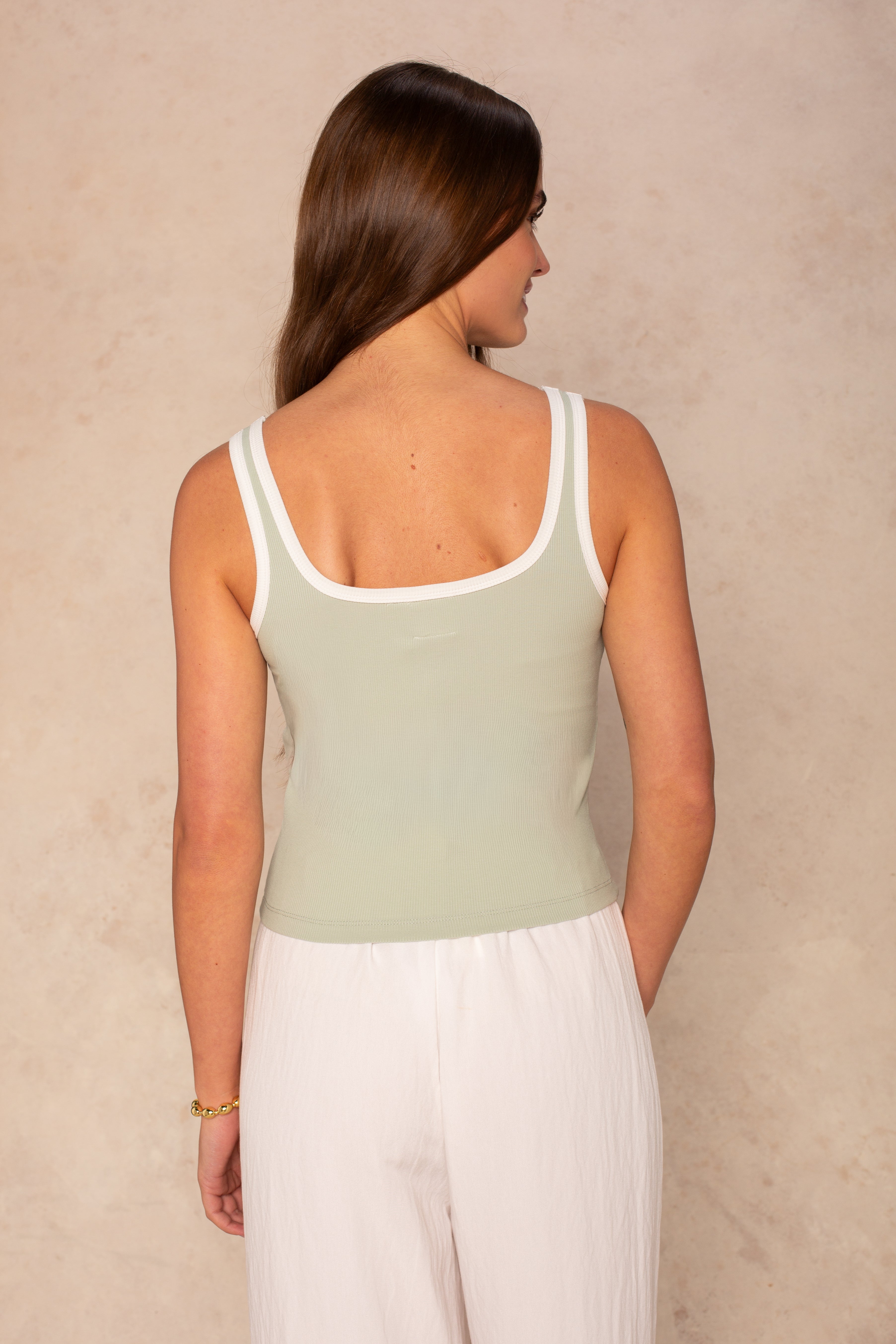 A woman with long brown hair faces away, wearing the Isla Maggie Two Tone Tank—a sleeveless, soft knit light green top with white trim—paired with loose white pants against a neutral textured background.