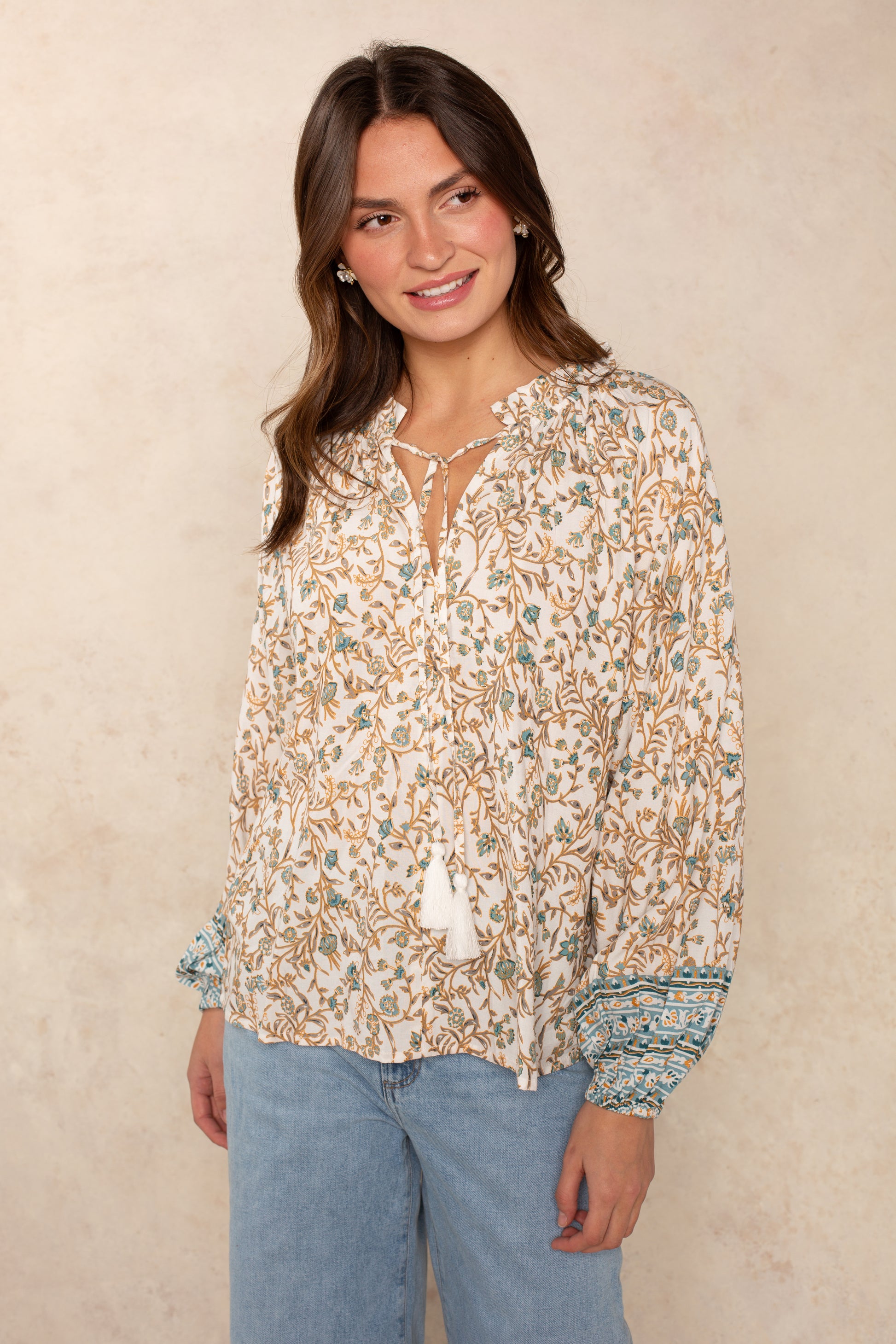 A woman models the Isla Whimsy Long Sleeve Woven Top in BLUE WHIMSY, featuring a floral print, ruffled neckline, tassel accents, and blue patterned cuffs. She pairs the flowy blouse with light blue jeans and poses against a beige background, her relaxed silhouette complemented by a gentle smile turned to the side.