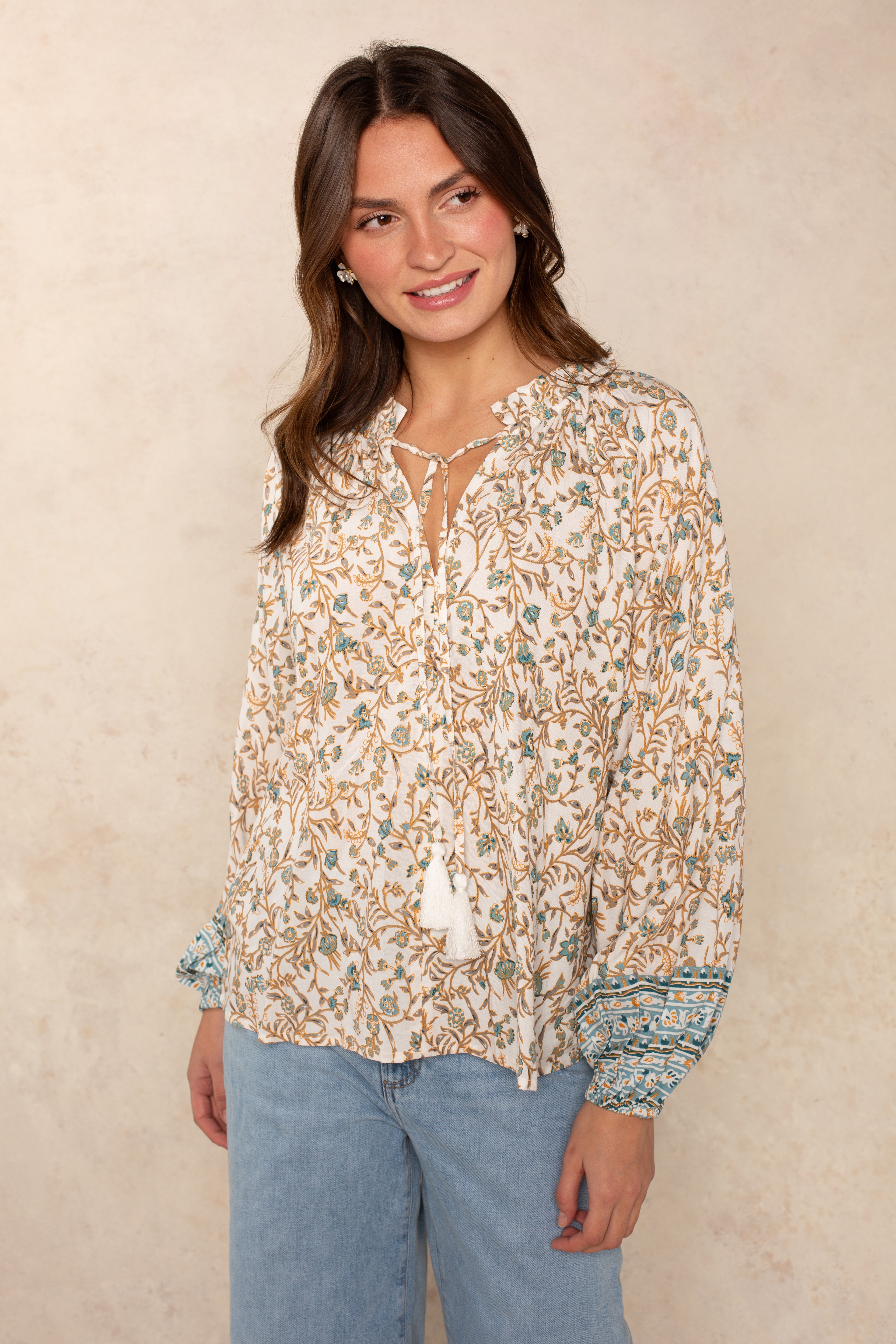 A woman models the Isla Whimsy Long Sleeve Woven Top in BLUE WHIMSY, featuring a floral print, ruffled neckline, tassel accents, and blue patterned cuffs. She pairs the flowy blouse with light blue jeans and poses against a beige background, her relaxed silhouette complemented by a gentle smile turned to the side.