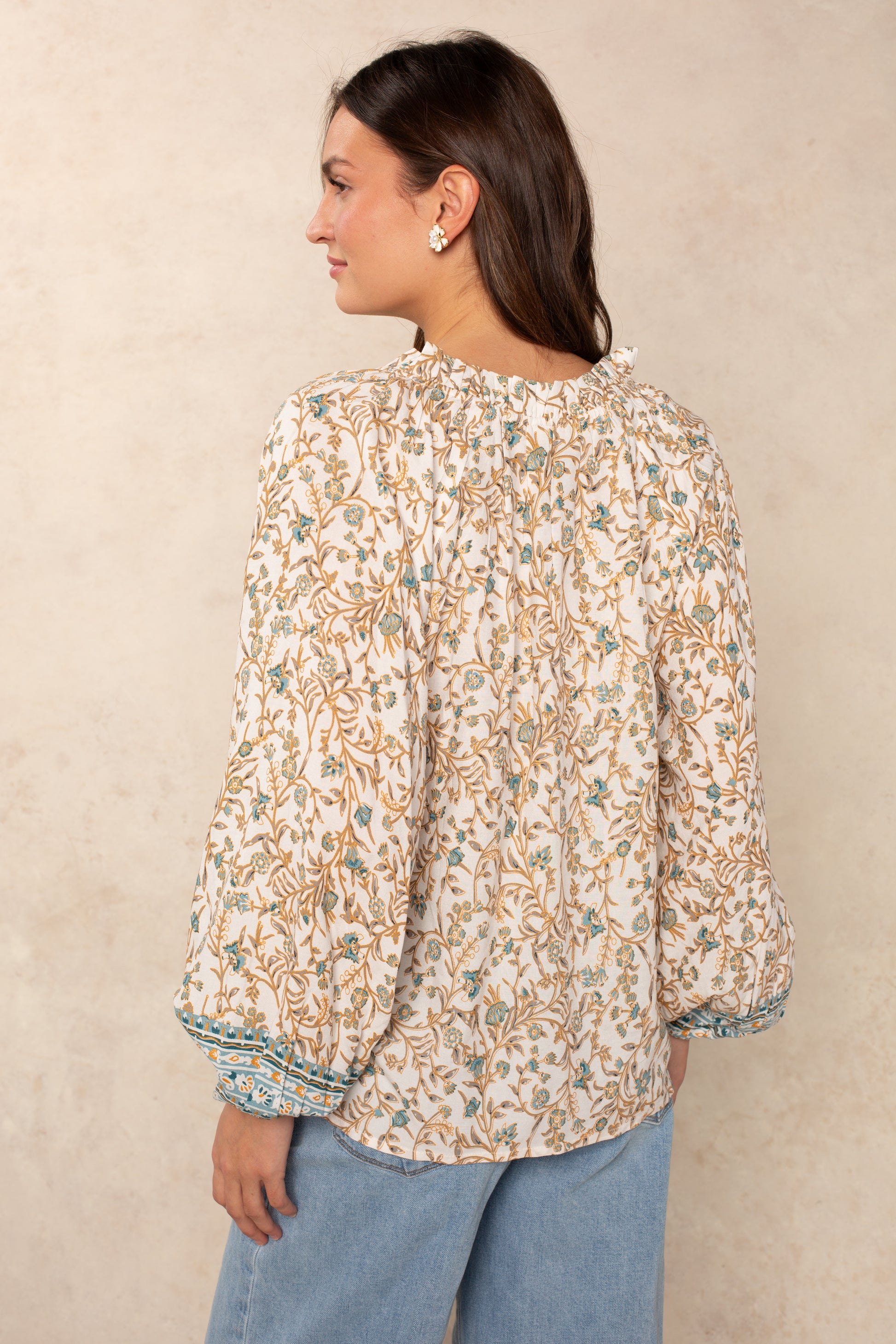 A woman with long brown hair wears the Isla Whimsy Long Sleeve Woven Top, which features a floral print, puffed sleeves, and a delicate ruffled neckline, paired with blue jeans. She faces away from the camera, highlighting the blouse’s back details and flowy silhouette against a neutral background.