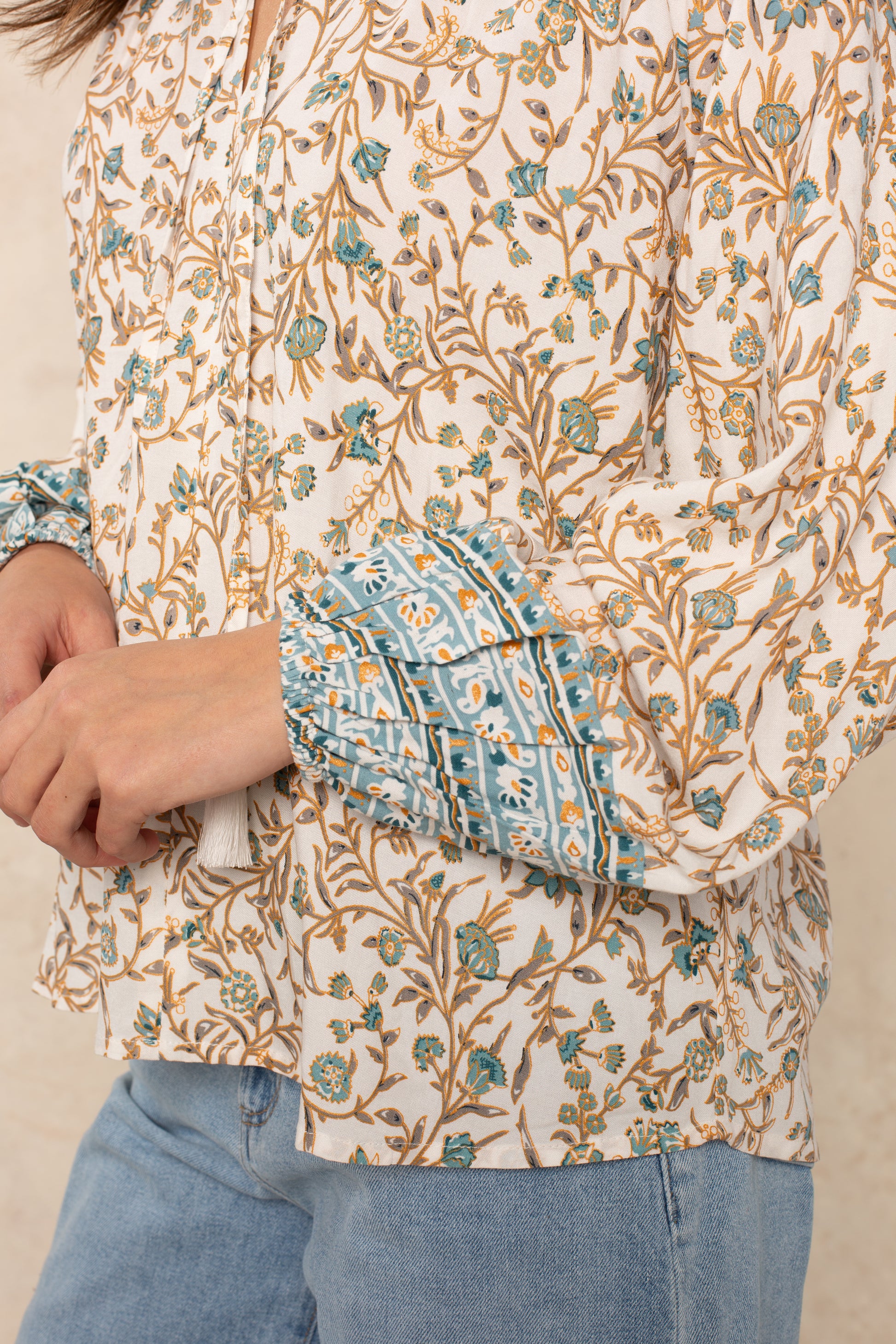 A person is wearing the Isla Whimsy Long Sleeve Woven Top—a floral print blouse with blue ornate cuffs, blouson sleeves, and a ruffled split neckline—paired with light blue jeans. Their arms are bent and only their torso and hands are visible.