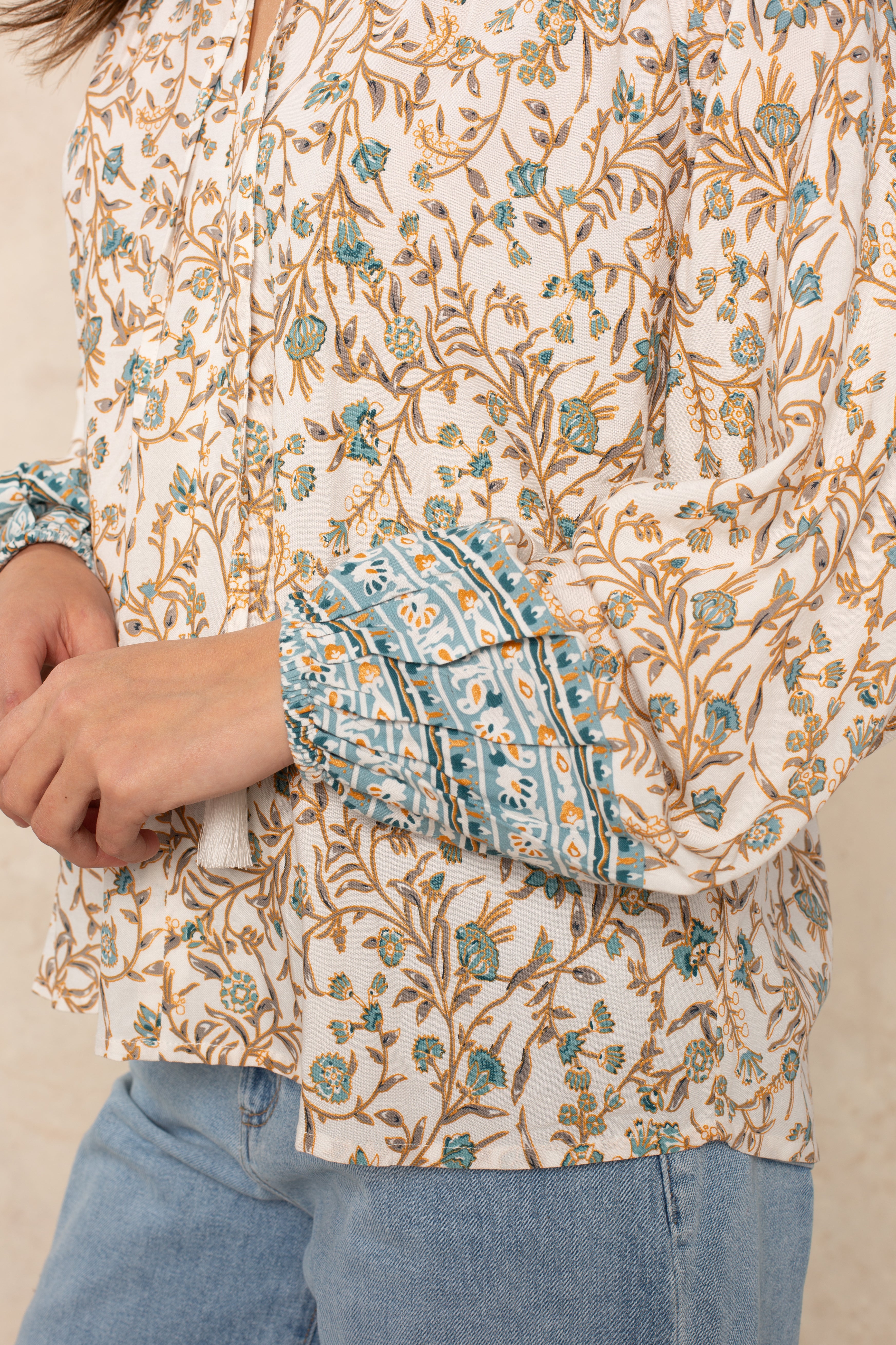 A person is wearing the Isla Whimsy Long Sleeve Woven Top—a floral print blouse with blue ornate cuffs, blouson sleeves, and a ruffled split neckline—paired with light blue jeans. Their arms are bent and only their torso and hands are visible.