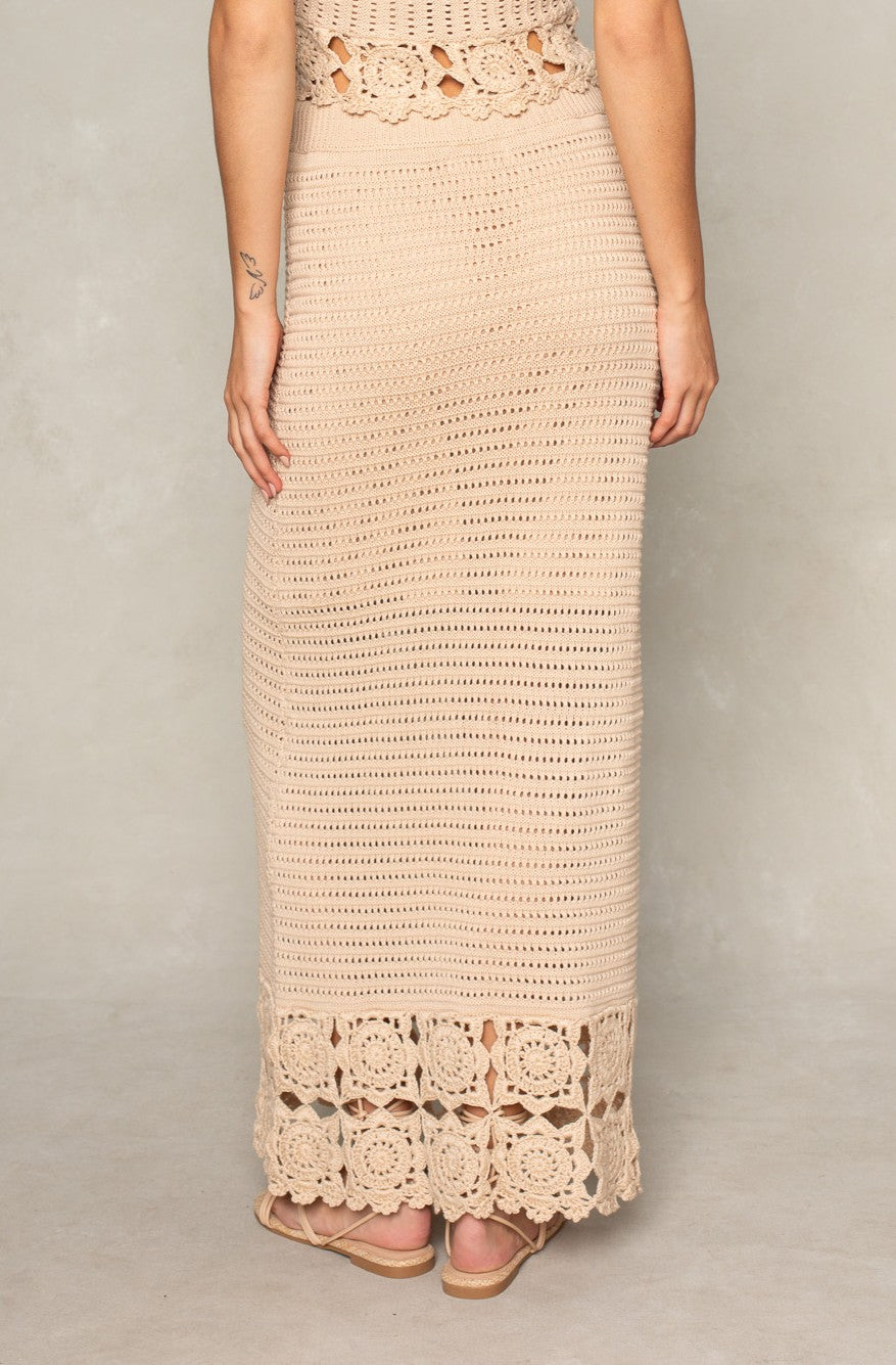 A person faces away, wearing Isla's Kai Crochet Maxi Skirt—a long, beige boho piece with floral crochet at the hem. Their left arm displays a small tattoo and they wear sandals, creating an effortless beachwear look.