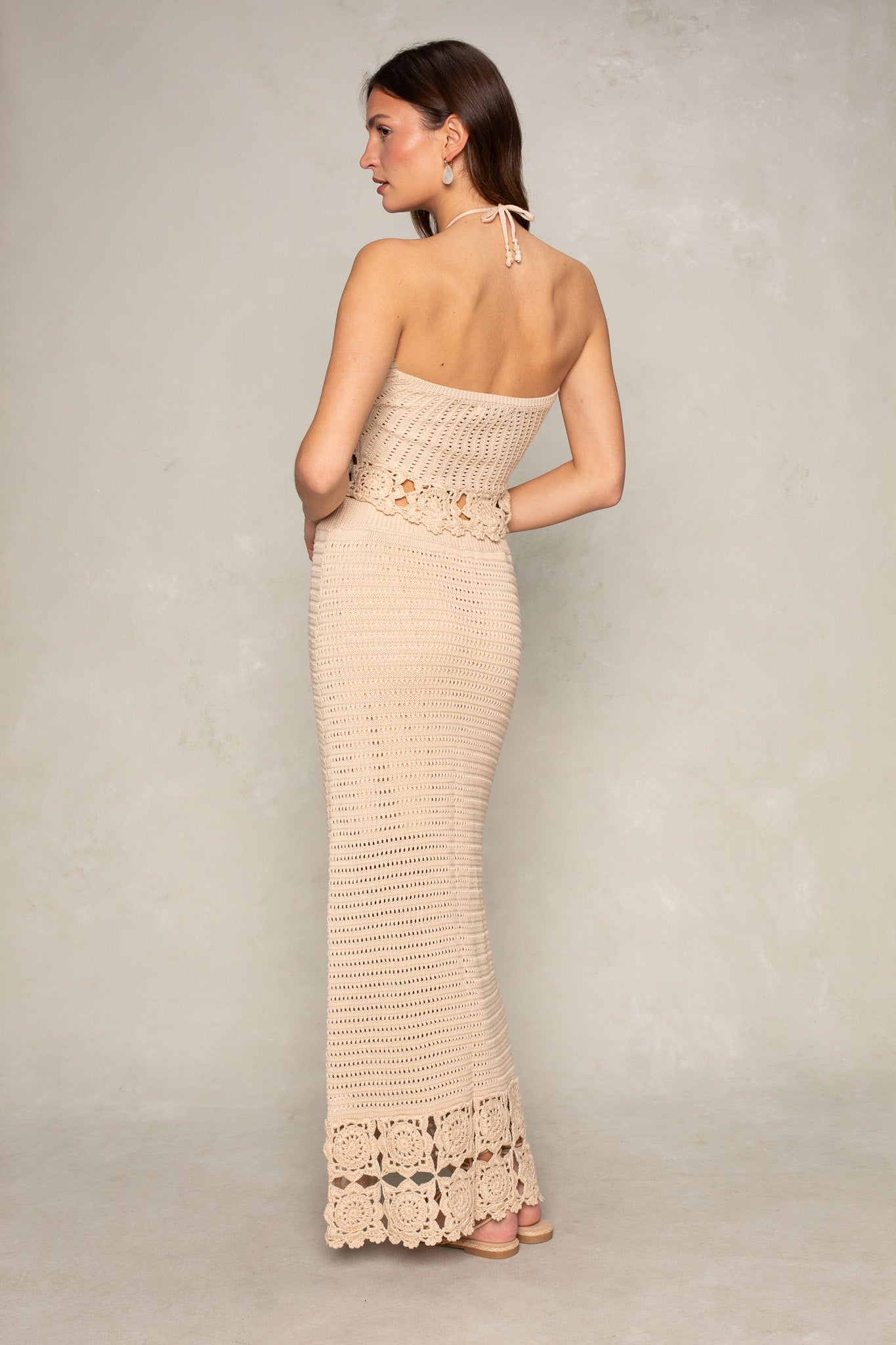A woman stands barefoot on a neutral background, wearing Isla’s beige Kai Crochet Maxi Skirt with floral crochet details at the waist and hem. She is turned to the side, looking over her shoulder.
