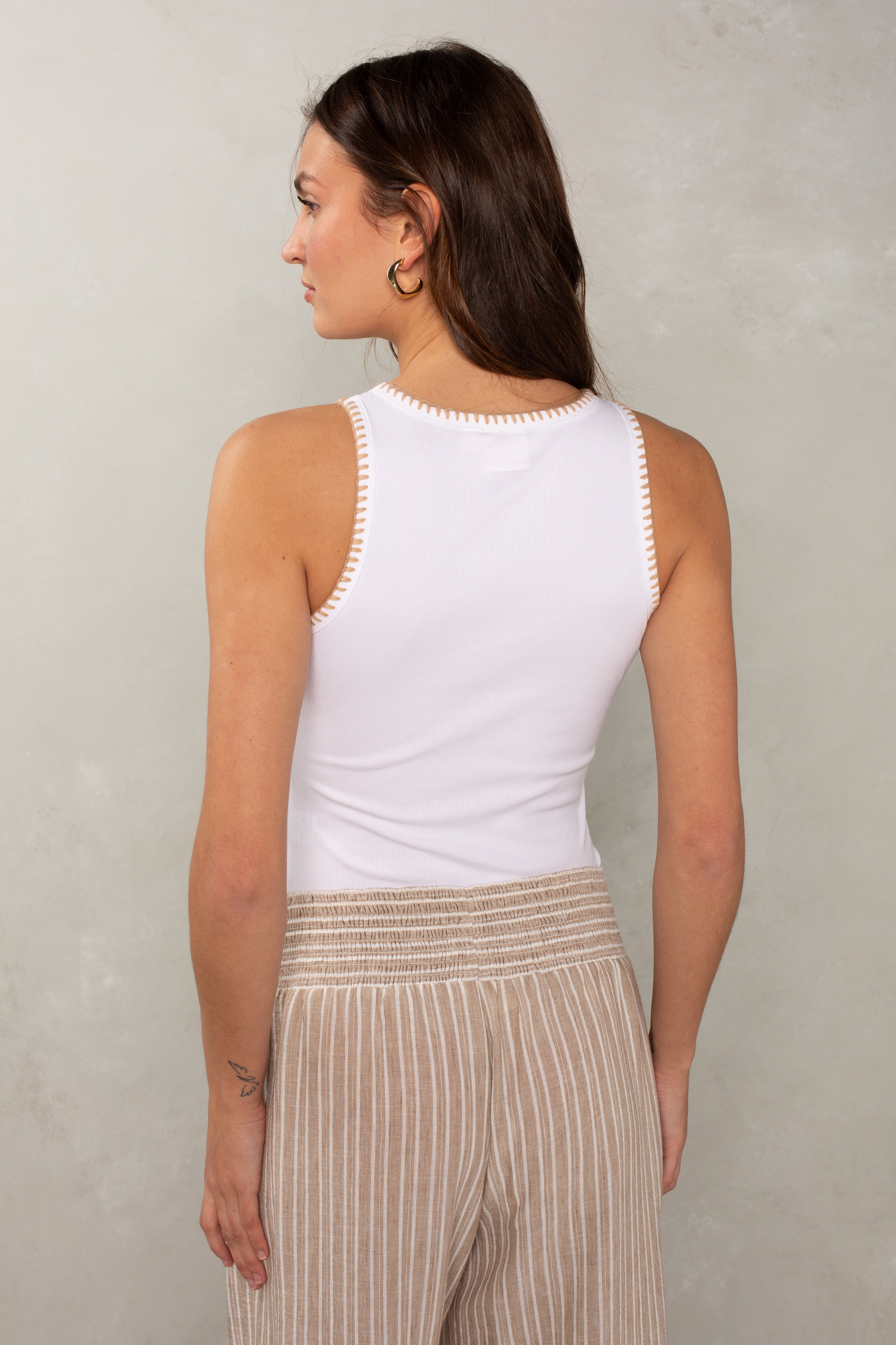 A woman with long brown hair is seen from the back wearing Isla’s Simone Stitch Tank in White, a lightweight sleeveless top, along with beige striped pants. She stands against a light gray textured background.