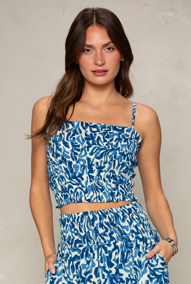 A woman with long brown hair wears the Isla Jules Smocked Cami in Blue Multi with adjustable ties and a matching skirt, standing against a plain light background with one hand in her pocket.