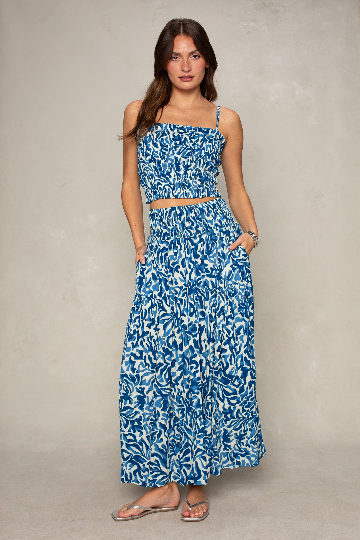 A woman stands against a neutral background wearing Isla’s Jules Smocked Skirt with pockets, paired with a matching sleeveless blue and white patterned crop top, and sandals.