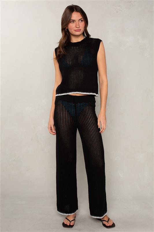 A woman with long brown hair stands against a light gray wall, wearing Isla's Eileen Crochet Coverup Pant in black over a blue bikini, paired with a sleeveless black crochet top and black sandals. The sheer wide-leg pants feature white trim.