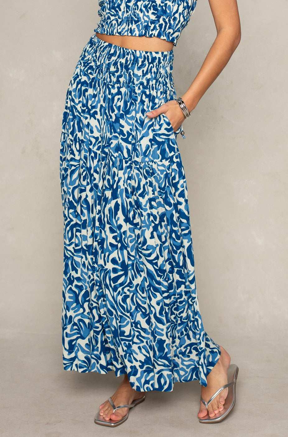 Wearing Isla's Jules Smocked Skirt in Blue Multi, this person pairs the versatile piece with a matching crop top, silver sandals and bracelets. They stand against a neutral background, hand in the skirt pocket to highlight its style.