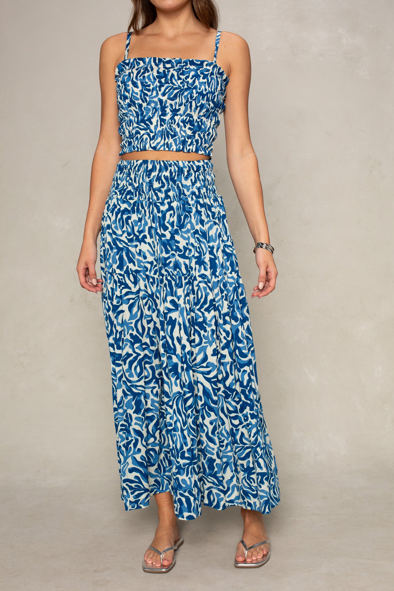 A person wearing the Isla Jules Smocked Cami in a blue and white leaf pattern, styled with a matching high-waisted maxi skirt and sandals, stands against a neutral background.