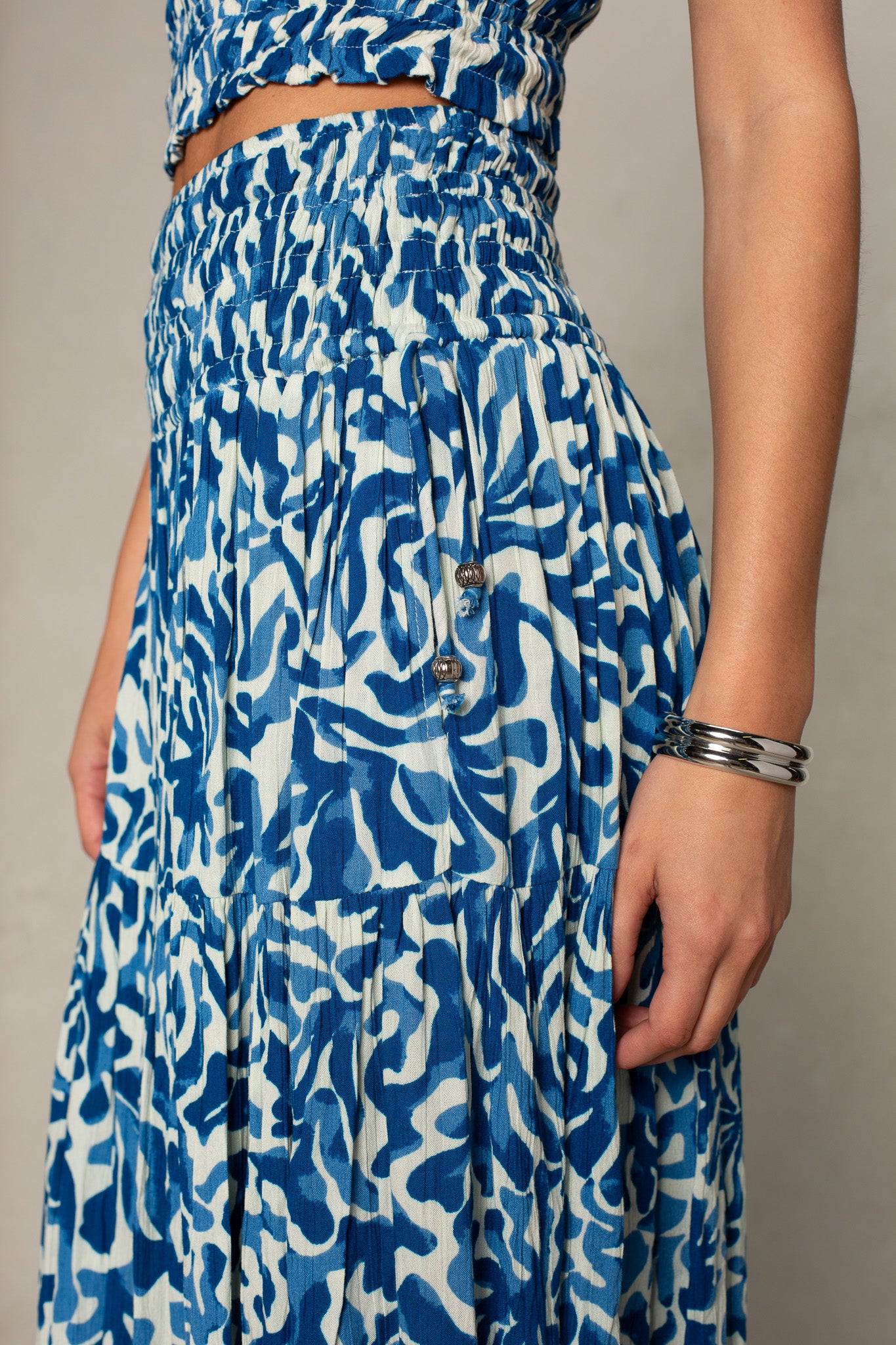 A person wears the Isla Jules Smocked Skirt, featuring a blue and white abstract pattern, pleats, smocking detail, and metal buttons. Silver bangles complete the look—this versatile skirt is perfect for layering.