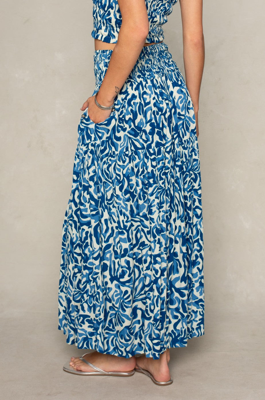 A person models the Isla Jules Smocked Skirt set—a blue and white patterned sleeveless top with smocking and a matching long skirt—sideways, hand in pocket, styled with silver sandals and bracelets against a neutral background.