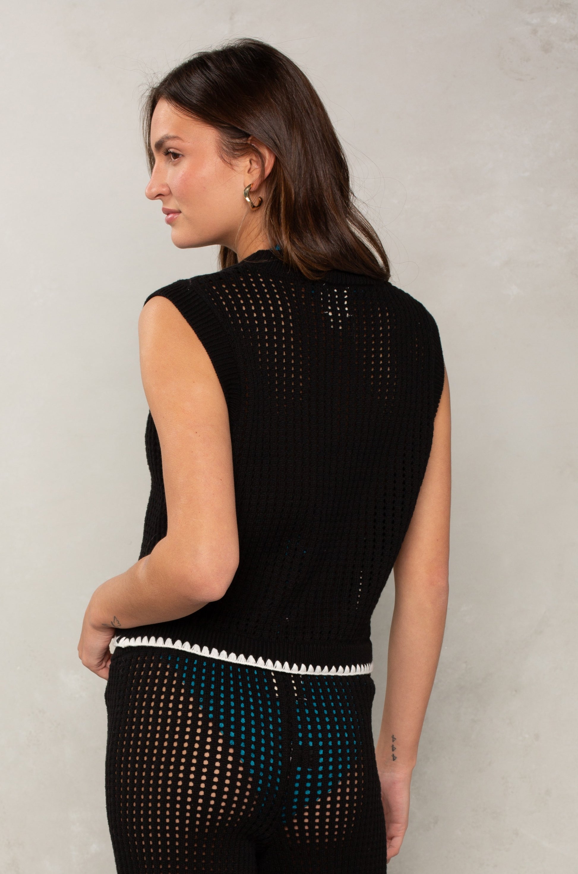 Woman facing away, wearing the Isla Eileen Crochet Coverup Top—a relaxed fit, open-knit sleeveless black crochet crop—paired with matching pants layered over teal undergarments, standing against a light gray textured wall.