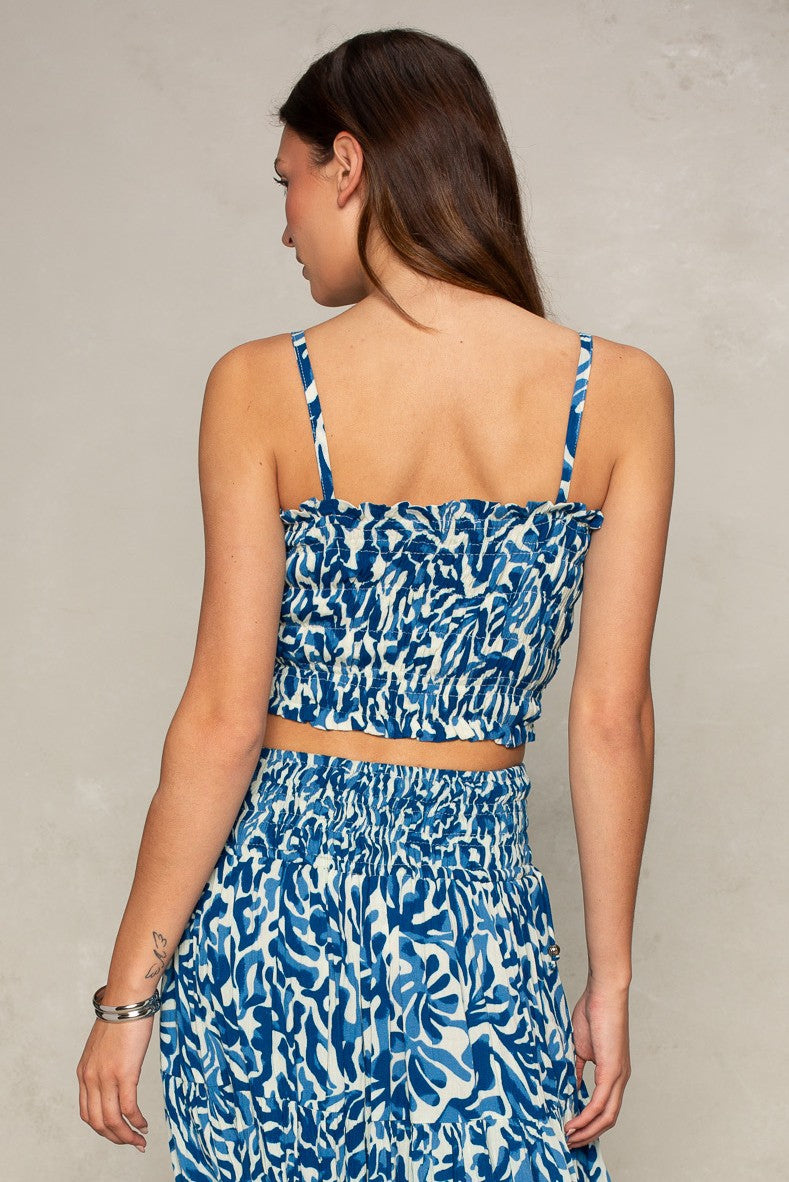 A woman with long brown hair faces away, wearing the Jules Smocked Cami by Isla in a blue and white pattern with matching skirt. The cropped cami features a smocked design and elastic waistband; she also wears a silver bracelet and shows a small left arm tattoo.