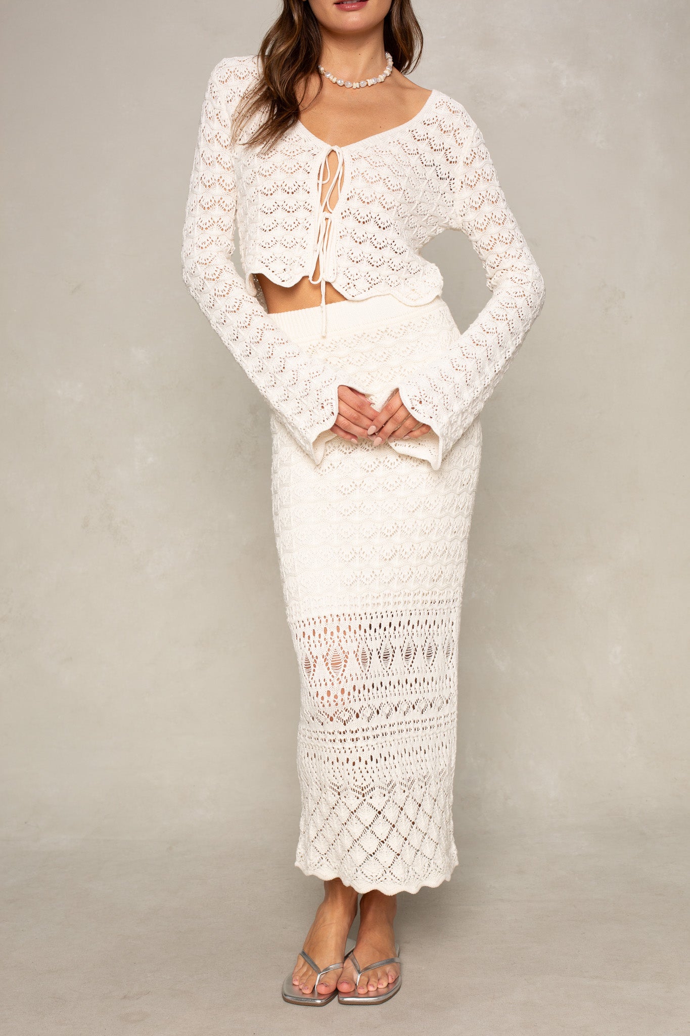 A woman in the Isla Andrea Two Tie Cardigan—a relaxed-fit, long-sleeved white top—with a matching high-waisted, ankle-length crochet skirt stands against a plain light background, accessorizing with sandals and a beaded necklace.