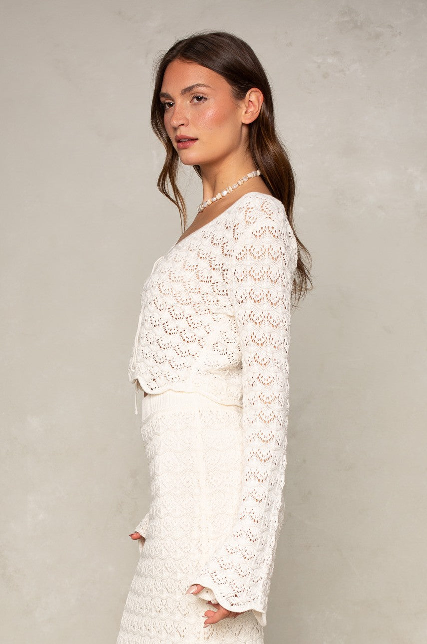A woman with long brown hair stands in profile, wearing the Isla Andrea Two Tie Cardigan—a relaxed fit white crocheted top—paired with a matching skirt and beaded necklace, set against a light textured background. Perfect for transitional seasons.