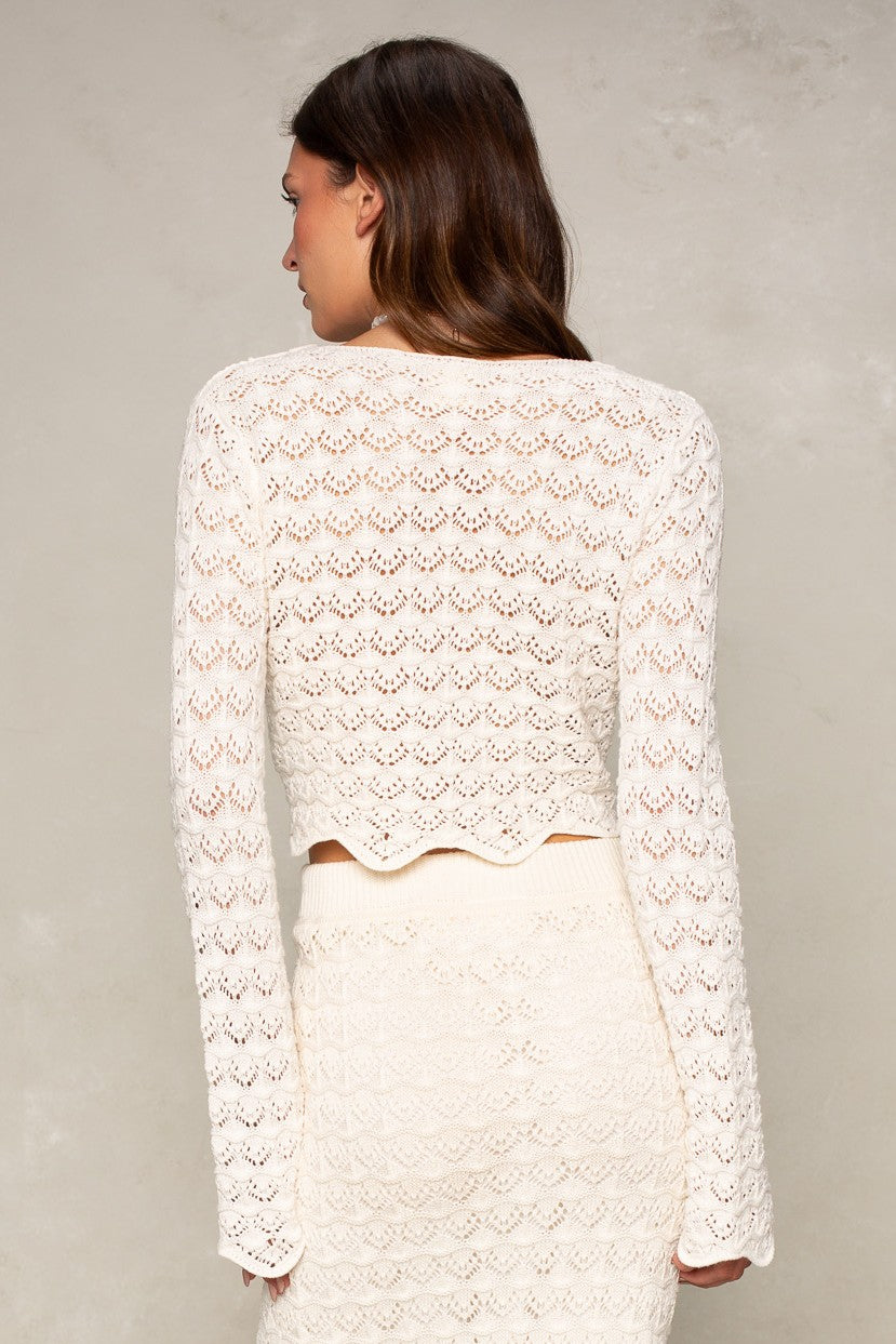 A woman with long brown hair faces away from the camera, wearing the Isla Andrea Two Tie Cardigan in a relaxed fit against a light textured background—an effortless look for transitional seasons.