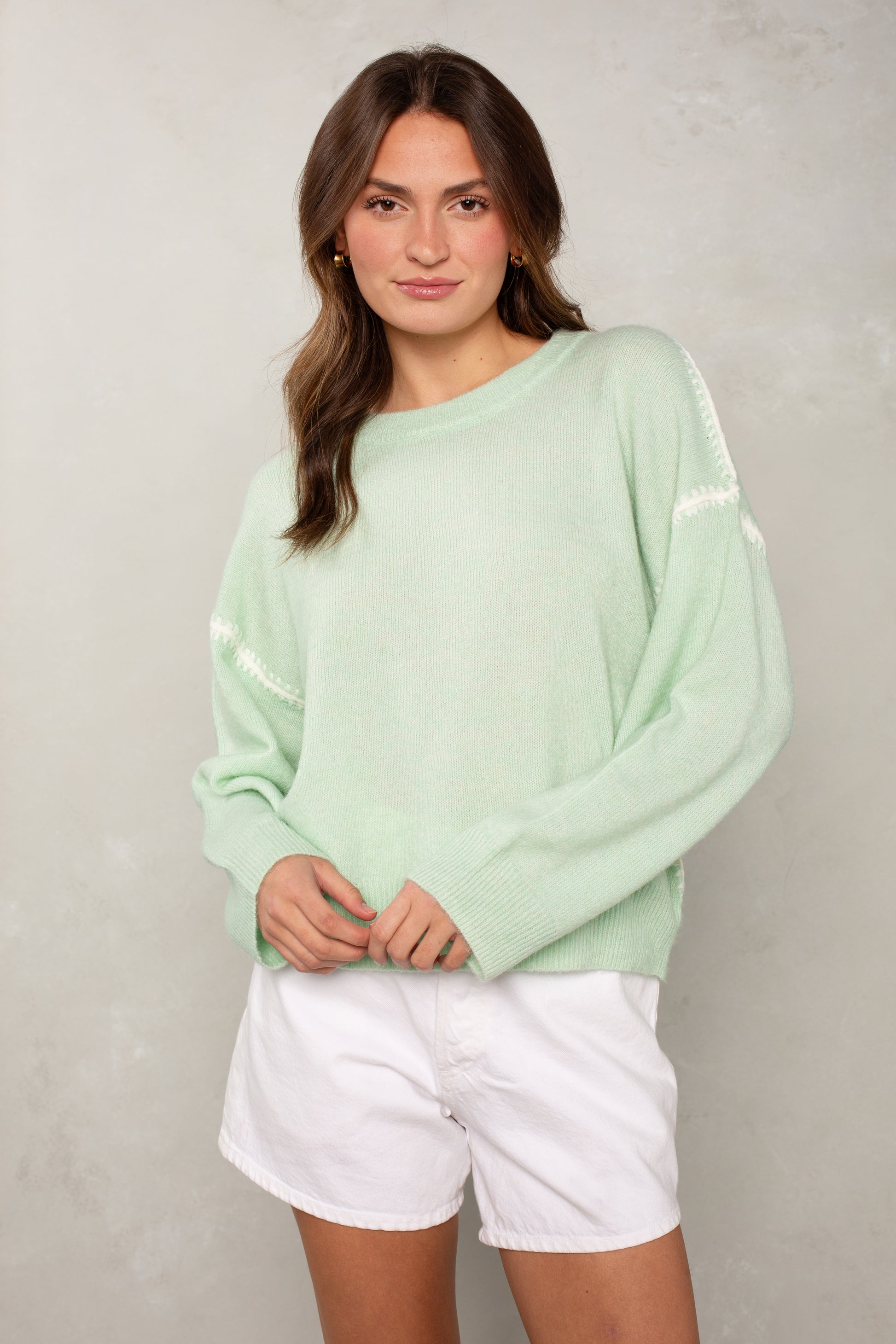 A woman smiles softly with her hands crossed at her waist, wearing Isla’s The Jackie Sweater in mint green with white detailing and subtle blanket stitching on the sleeves, paired with white shorts, against a light gray textured background.
