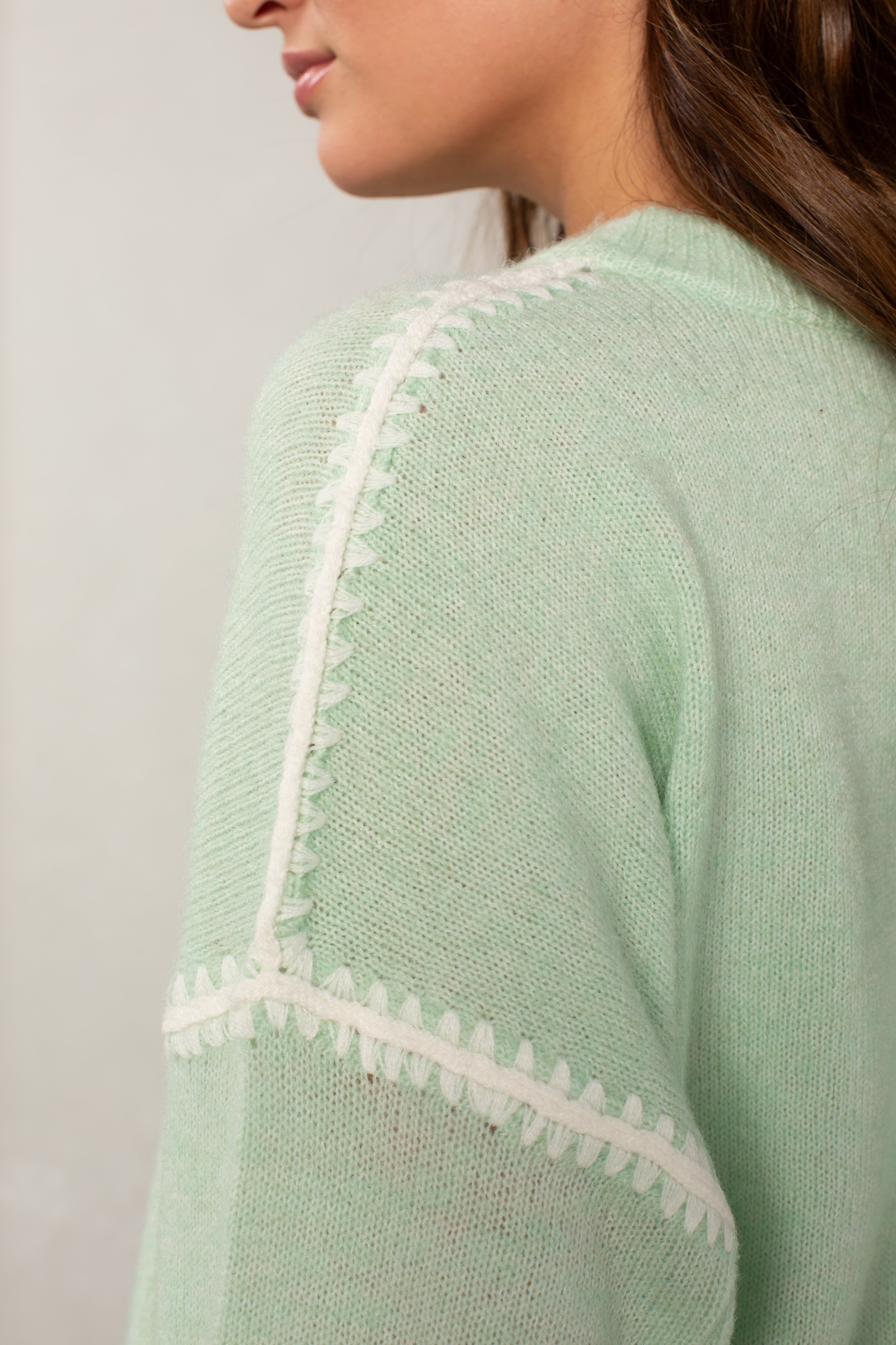 A close-up shows a woman wearing The Jackie Sweater by Isla, an oversized mint green knit with white blanket stitching on the shoulder and sleeve. Only her lower face, neck, and hair are partially visible.