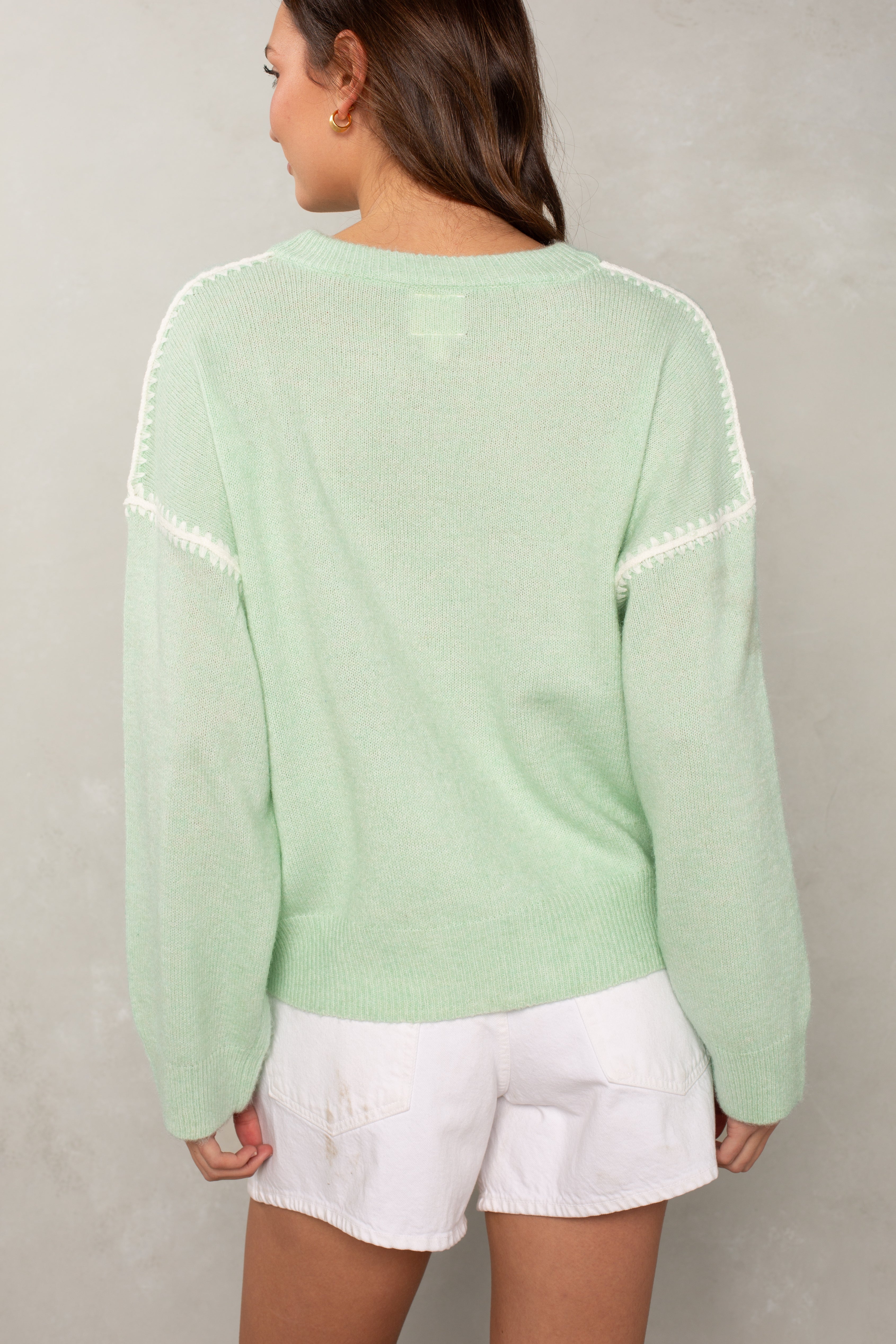 A woman with long brown hair stands with her back to the camera, wearing Isla’s The Jackie Sweater in mint green with blanket stitching and white shorts. The oversized sweater is set against a plain, light-colored wall.
