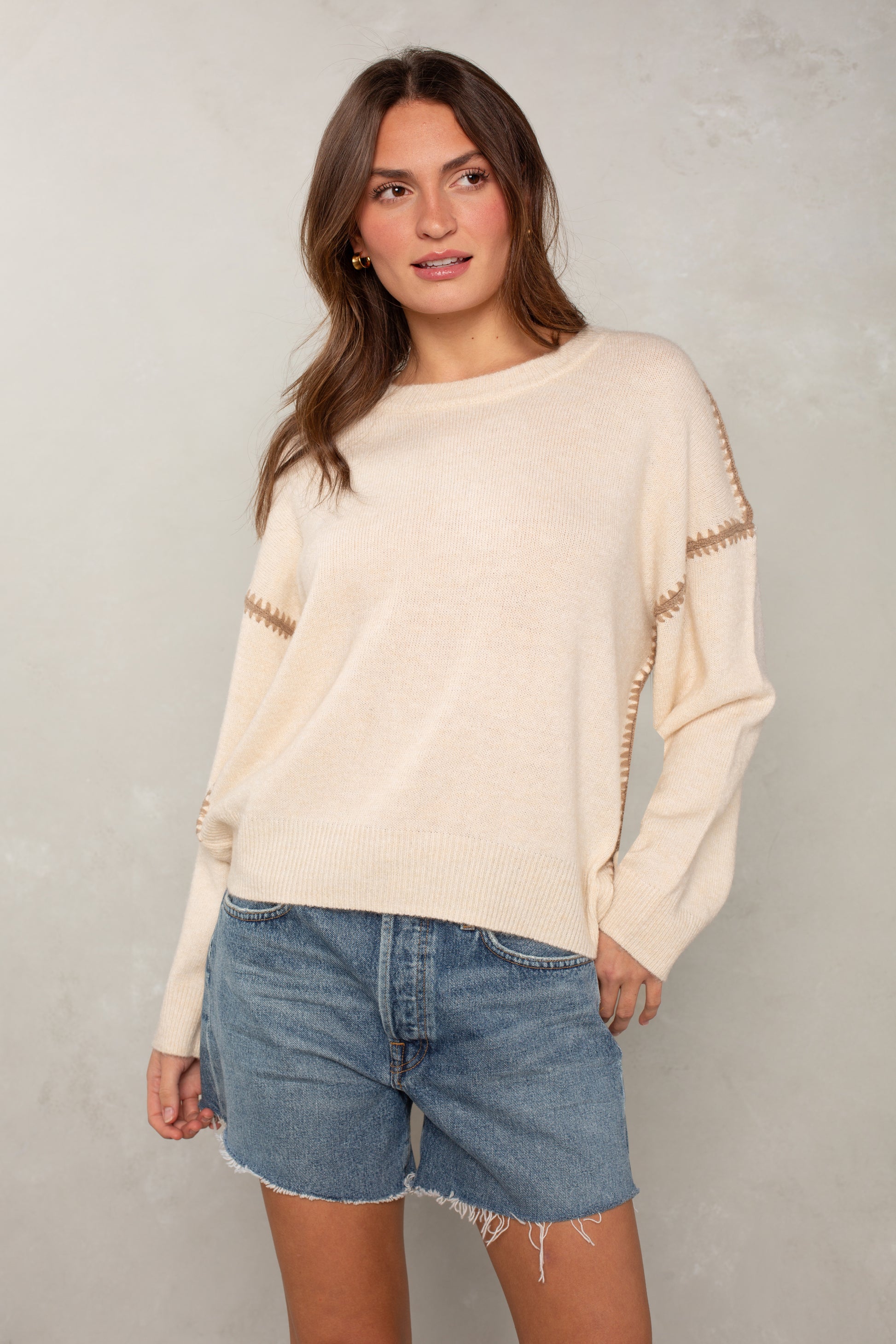 A woman with long brown hair stands against a light textured background wearing Isla’s The Jackie Sweater—an oversized cream sweater with brown blanket stitching—and blue denim shorts. She looks to the side, one hand in her pocket.