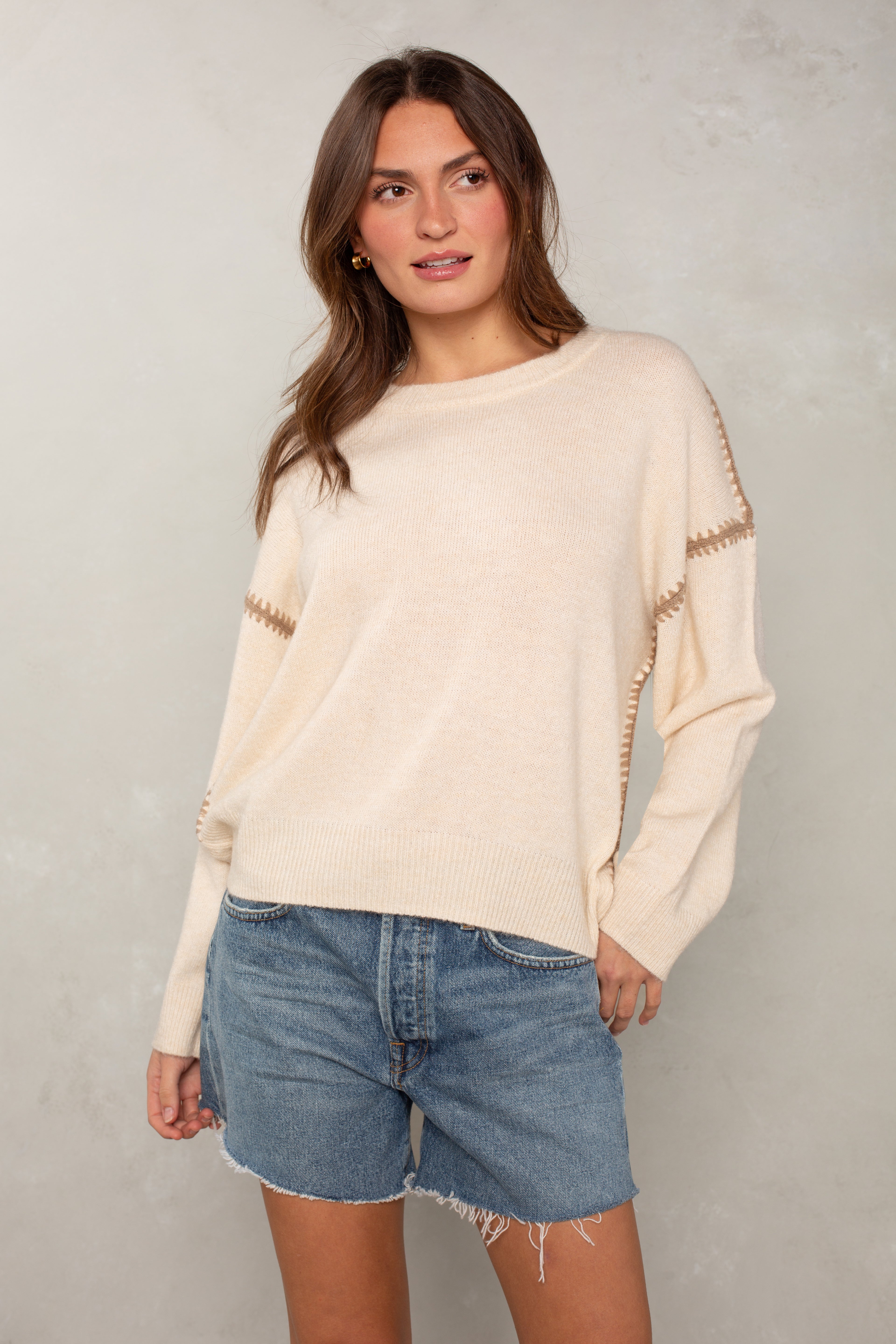 A woman with long brown hair stands against a light textured background wearing Isla’s The Jackie Sweater—an oversized cream sweater with brown blanket stitching—and blue denim shorts. She looks to the side, one hand in her pocket.