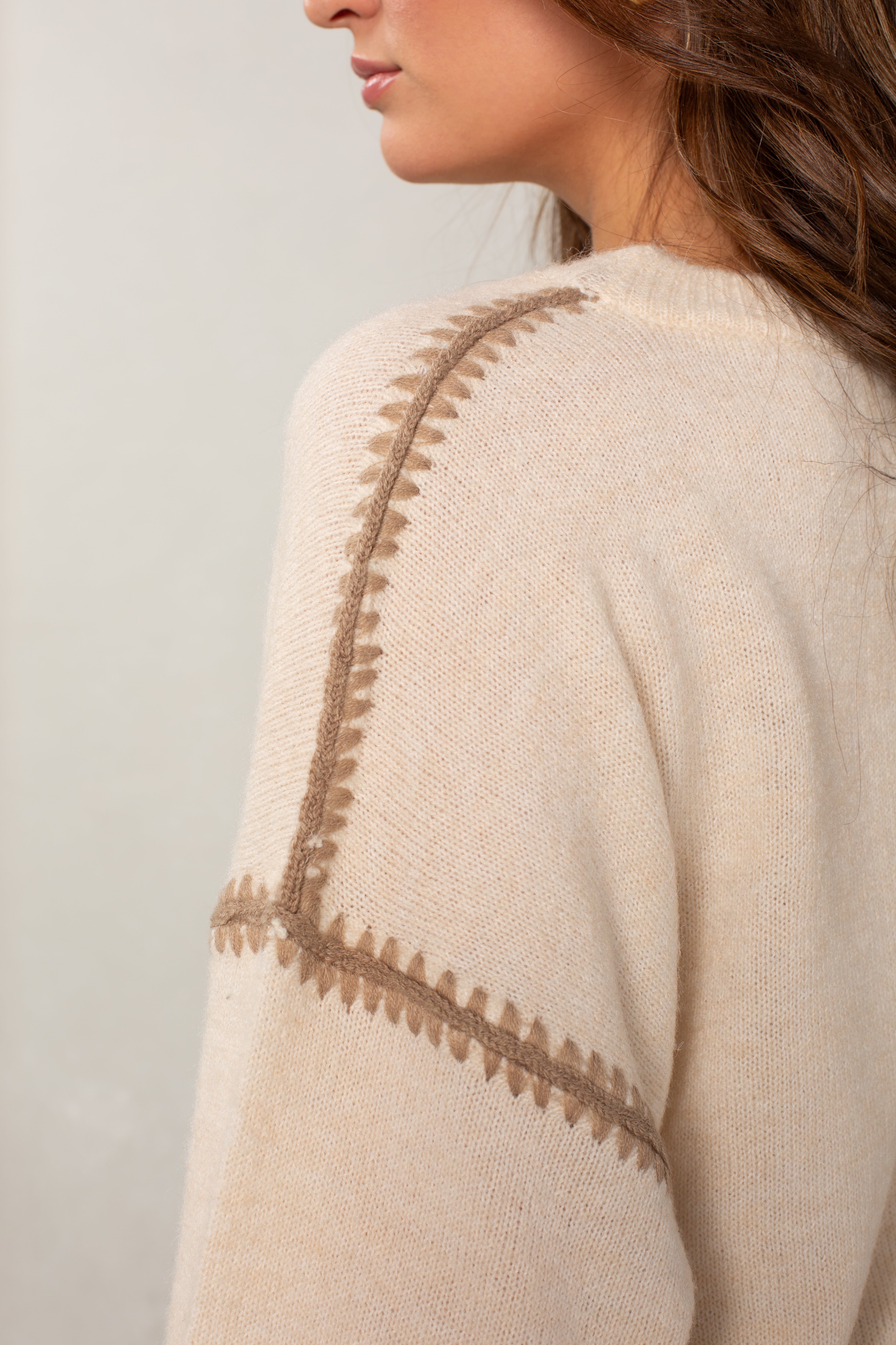 A close-up side view of a woman in Isla’s The Jackie Sweater, a cream-colored, oversized piece with light brown blanket stitching on the shoulder and sleeve seam. Her loosely wavy brown hair is partially visible.