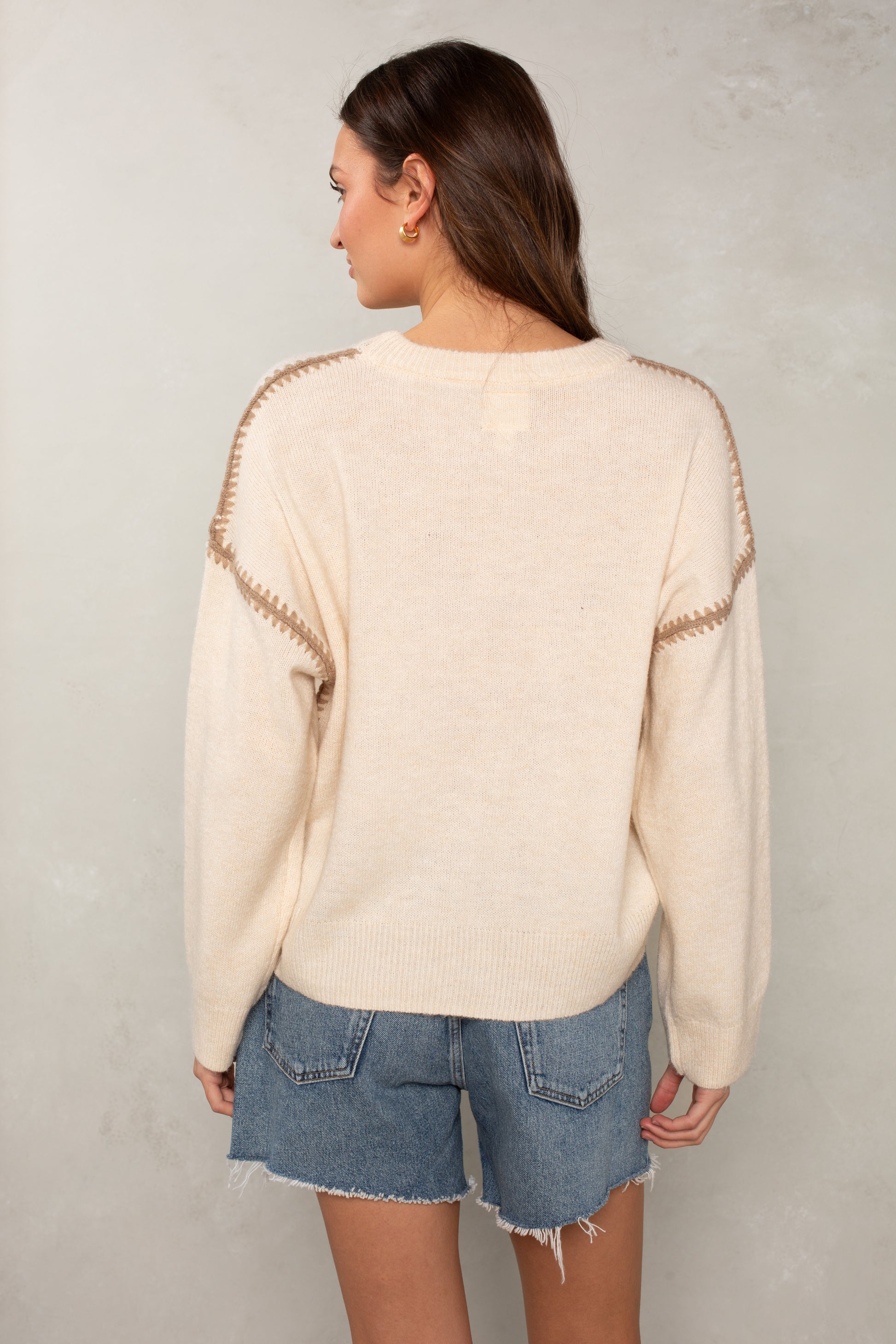 A woman with long brown hair stands with her back to the camera, wearing Isla's The Jackie Sweater in cream with blanket-stitched shoulders and blue frayed-hem denim shorts, against a light textured wall.