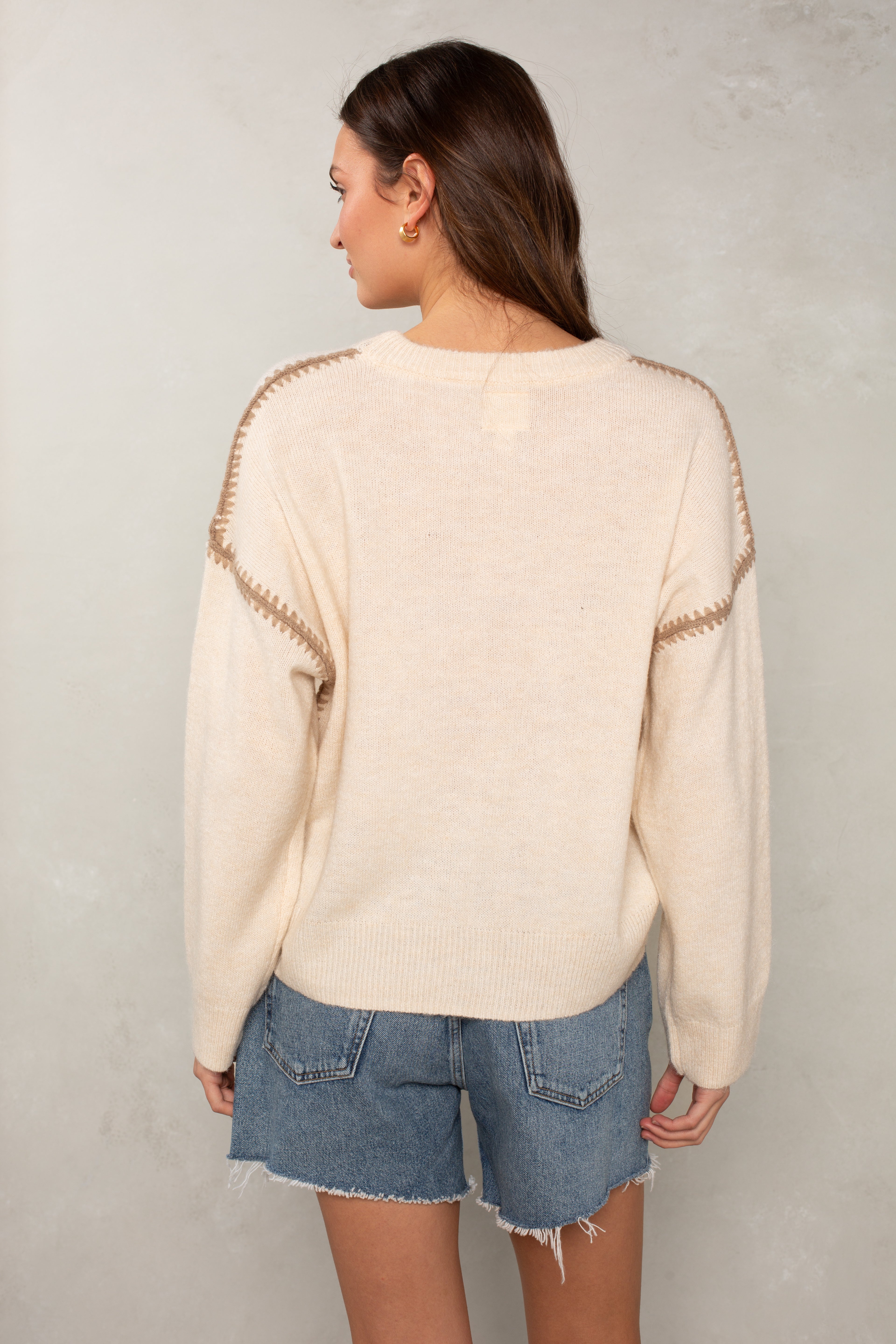 A woman with long brown hair stands with her back to the camera, wearing Isla's The Jackie Sweater in cream with blanket-stitched shoulders and blue frayed-hem denim shorts, against a light textured wall.