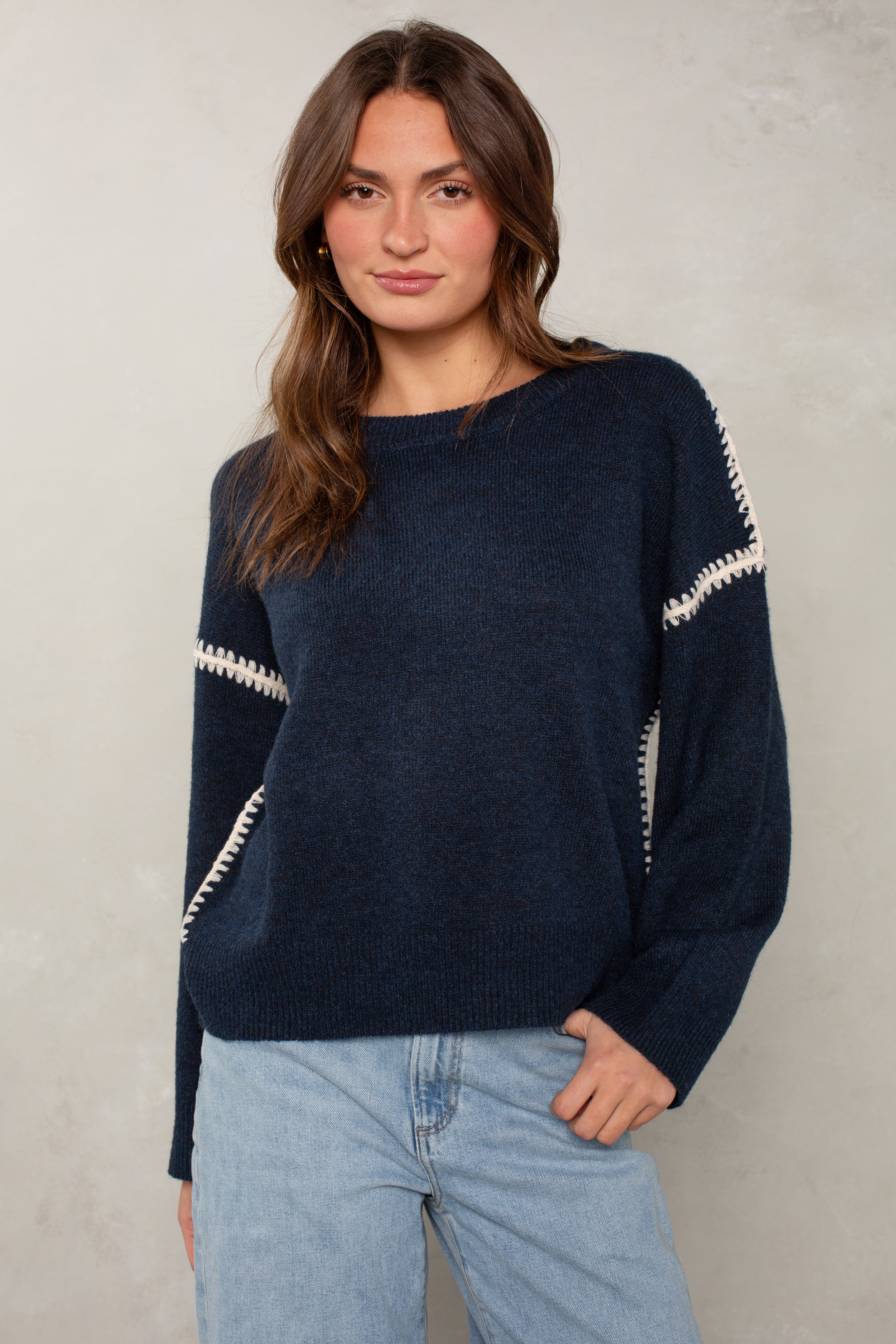 A woman with long brown hair wears The Jackie Sweater by Isla—a navy blue sweater with blanket stitching on the sleeves—and light blue jeans, standing against a light gray background and looking at the camera with a relaxed expression.