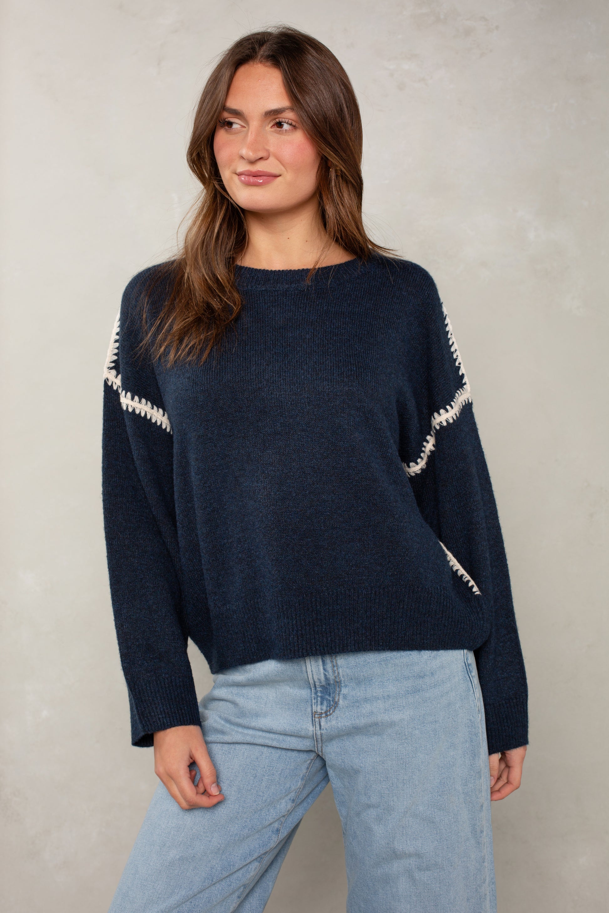 A woman with long brown hair models Isla's The Jackie Sweater—an oversized navy knit featuring white zigzag details and blanket stitching on the sleeves—styled with light blue jeans against a pale textured backdrop.